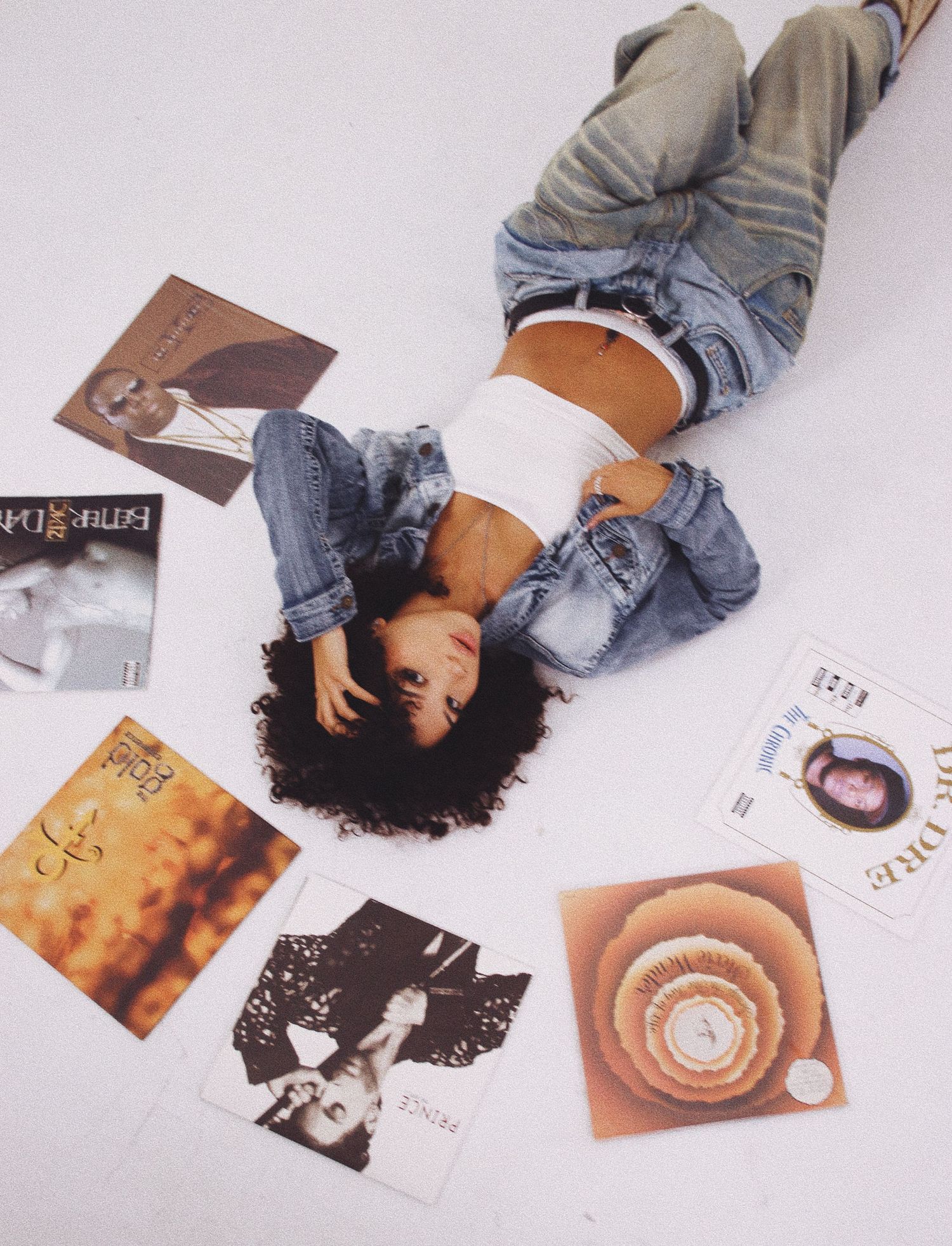 Person lying on white floor surrounded by vinyl records and album covers in casual denim outfit.