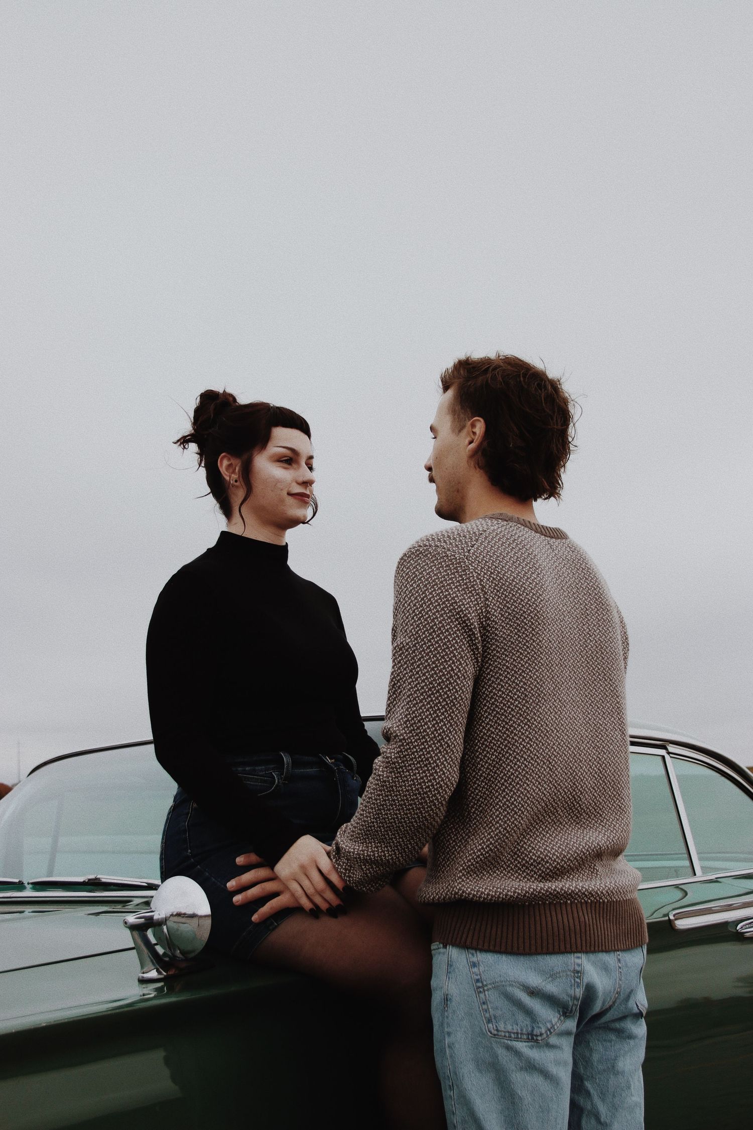 A couple leans against a vintage green car, sharing a tender moment under an overcast sky.