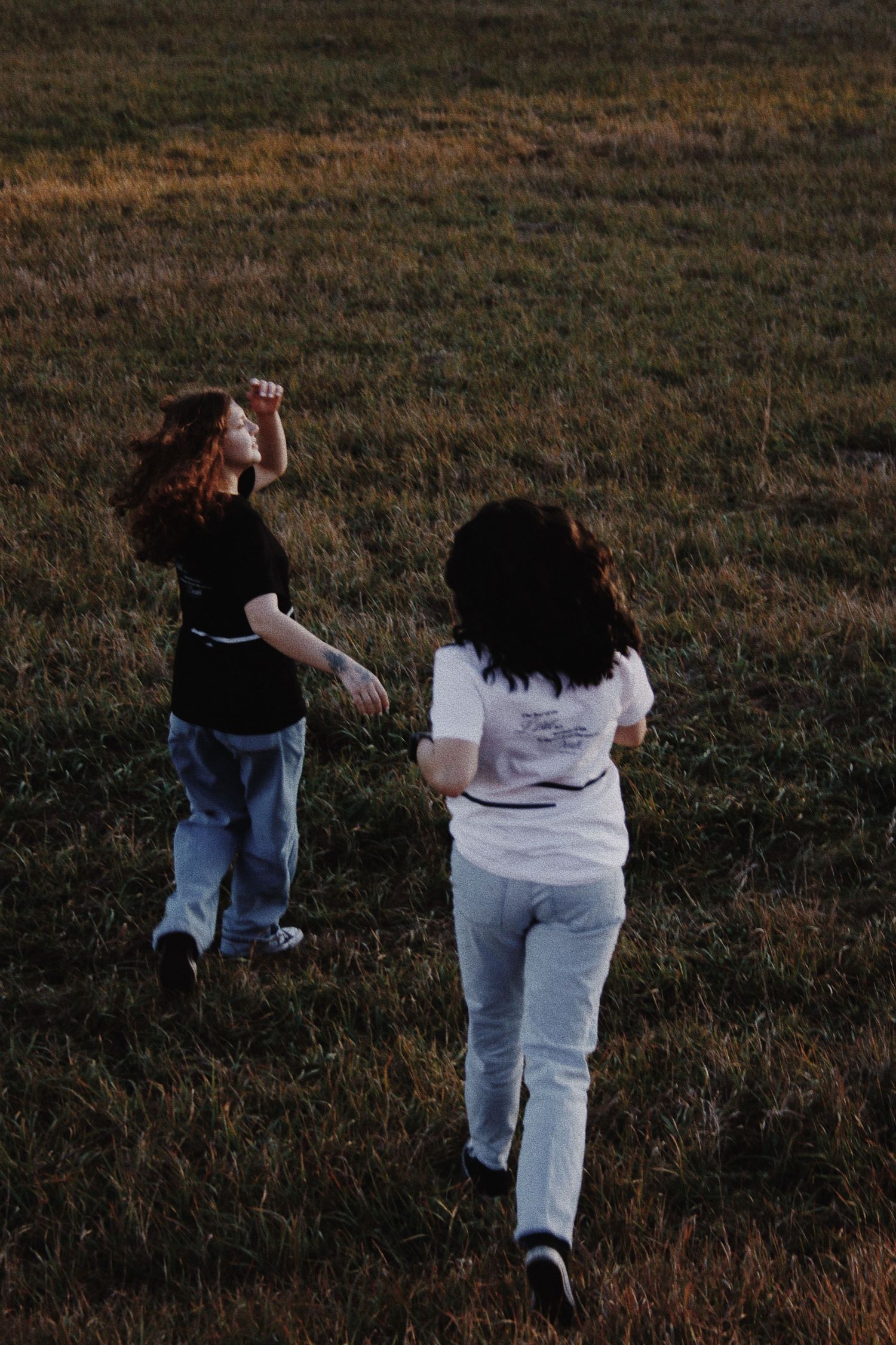 Two girls running through a grassy field at dusk, one in black tee and jeans, other in white tee and grey pants.