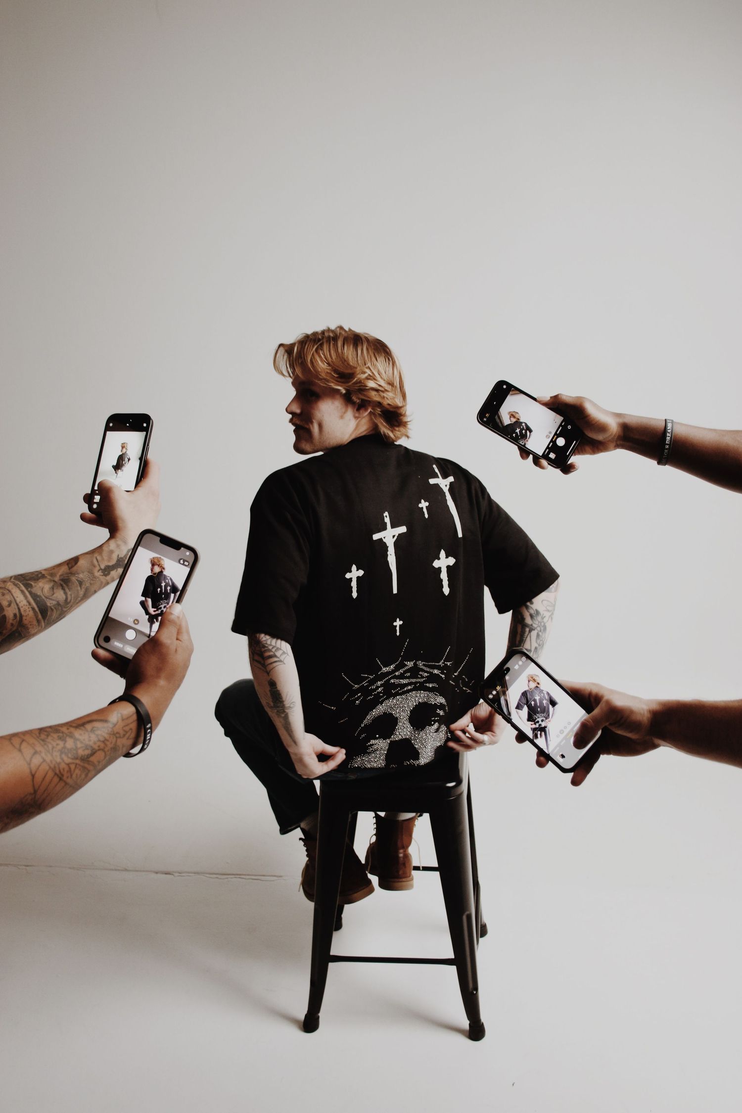 Man in black graphic cross t-shirt sits on stool surrounded by hands holding smartphones photographing him.