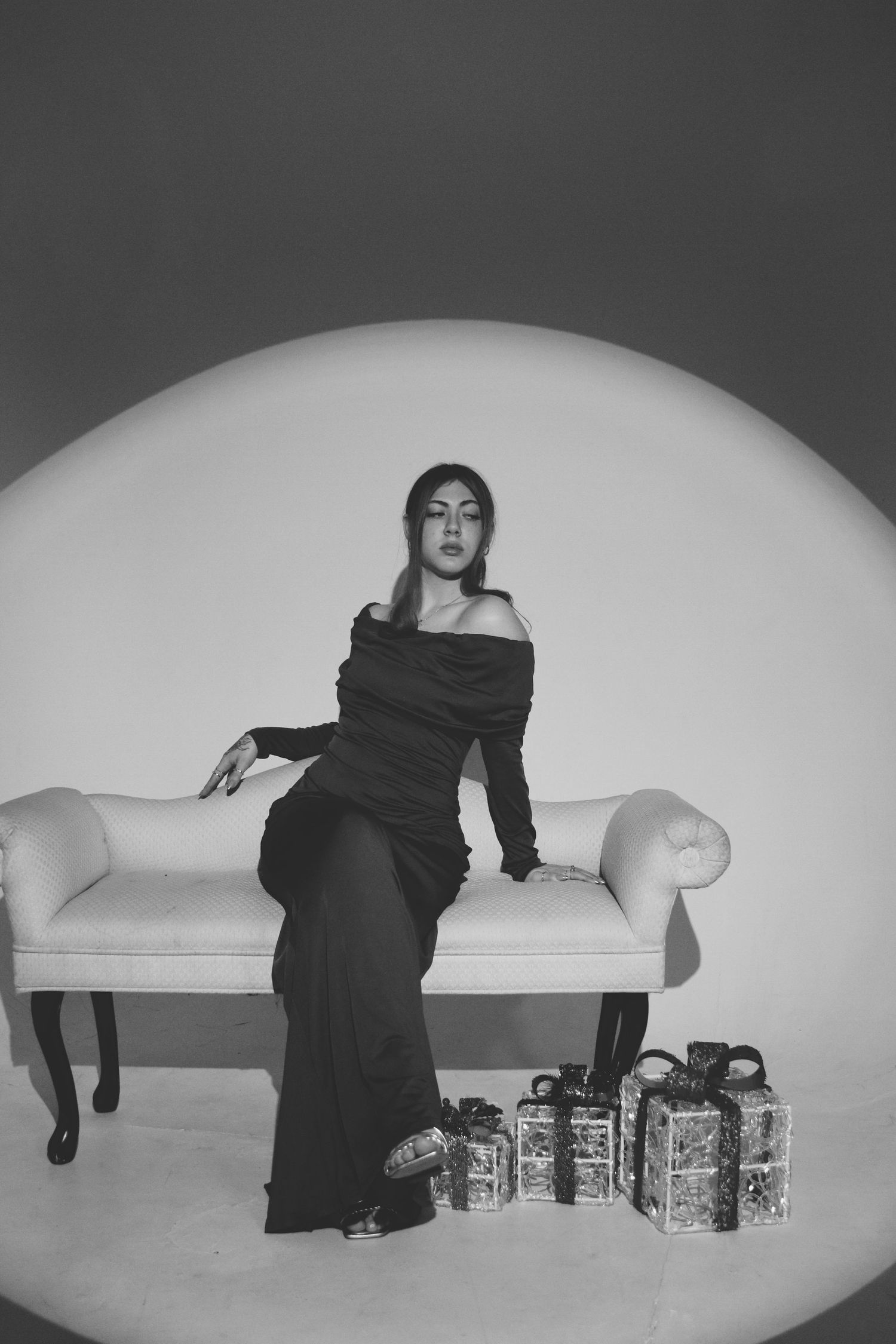 Elegant woman in black off-shoulder gown poses on white chaise with gift boxes in B&W photo.