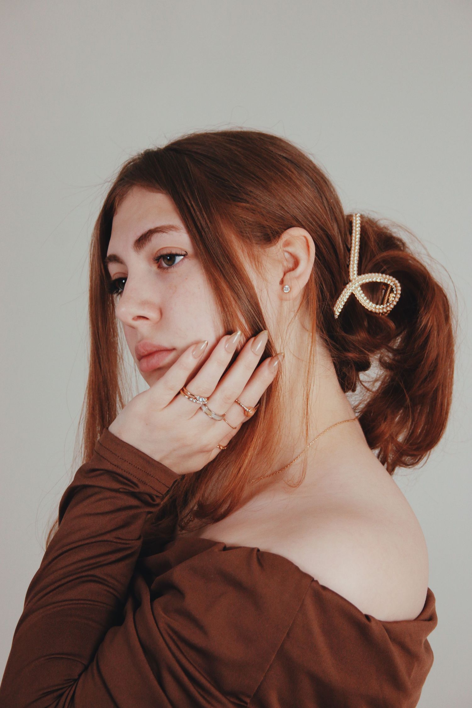 Young woman with auburn hair in pearl clip, wearing gold rings and off-shoulder brown top, posing against gray background.