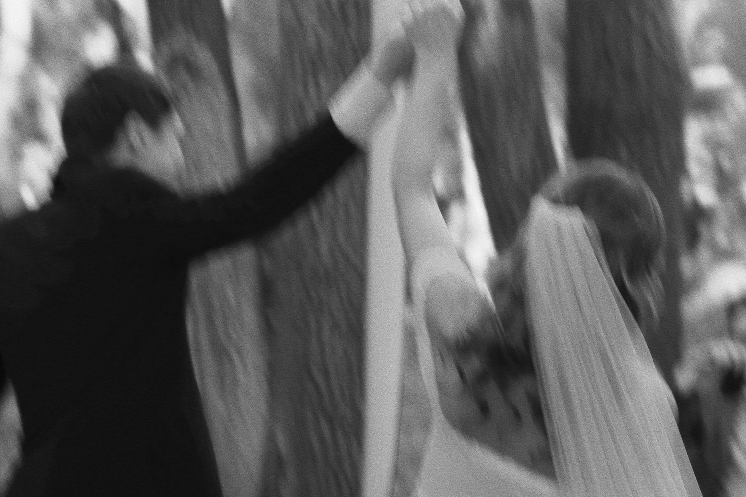 Black and white photo of a bride and groom dancing outdoors among trees on their wedding day.