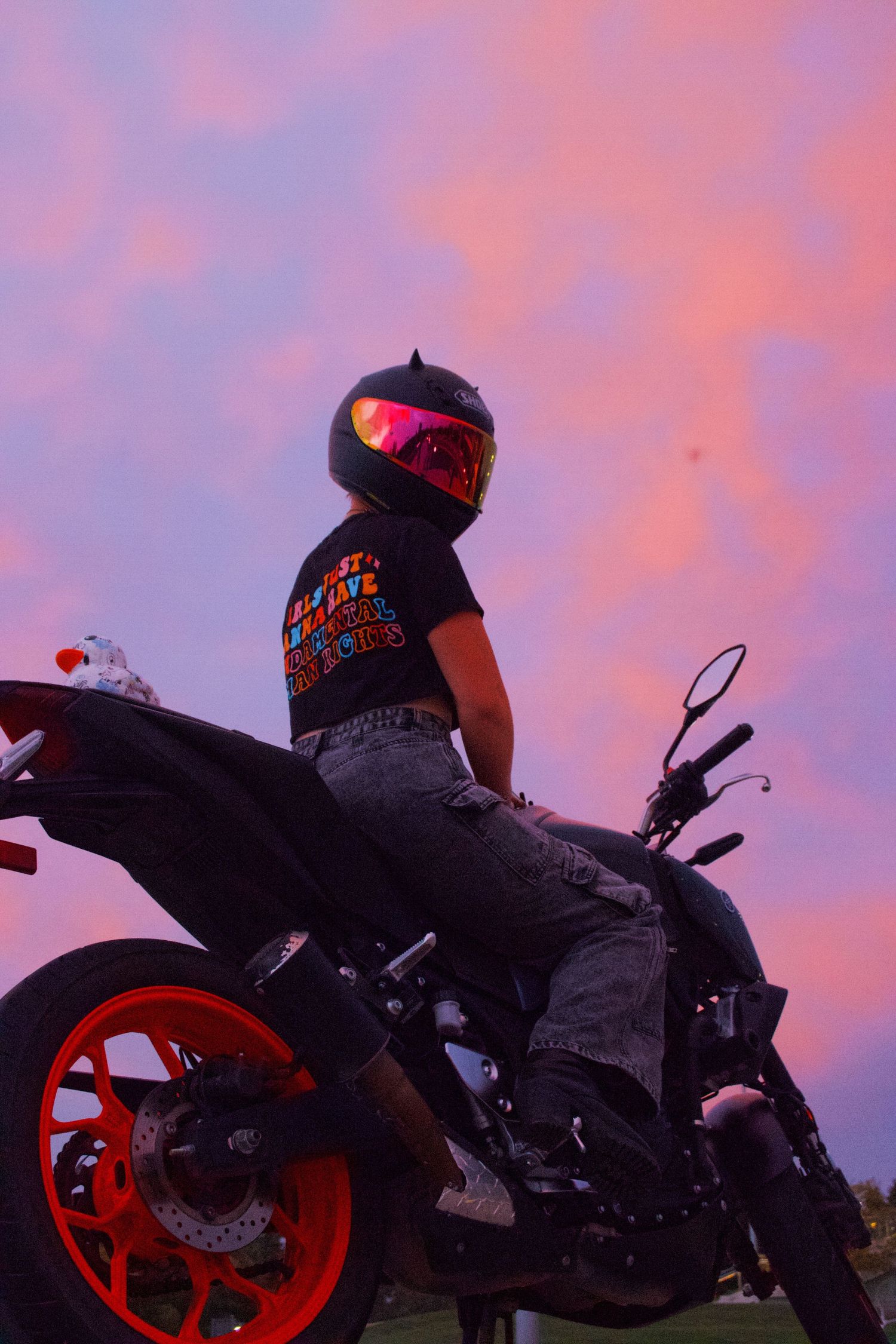 Motorcyclist in black helmet and t-shirt sits on red and black sport bike against vibrant pink and purple sunset sky.