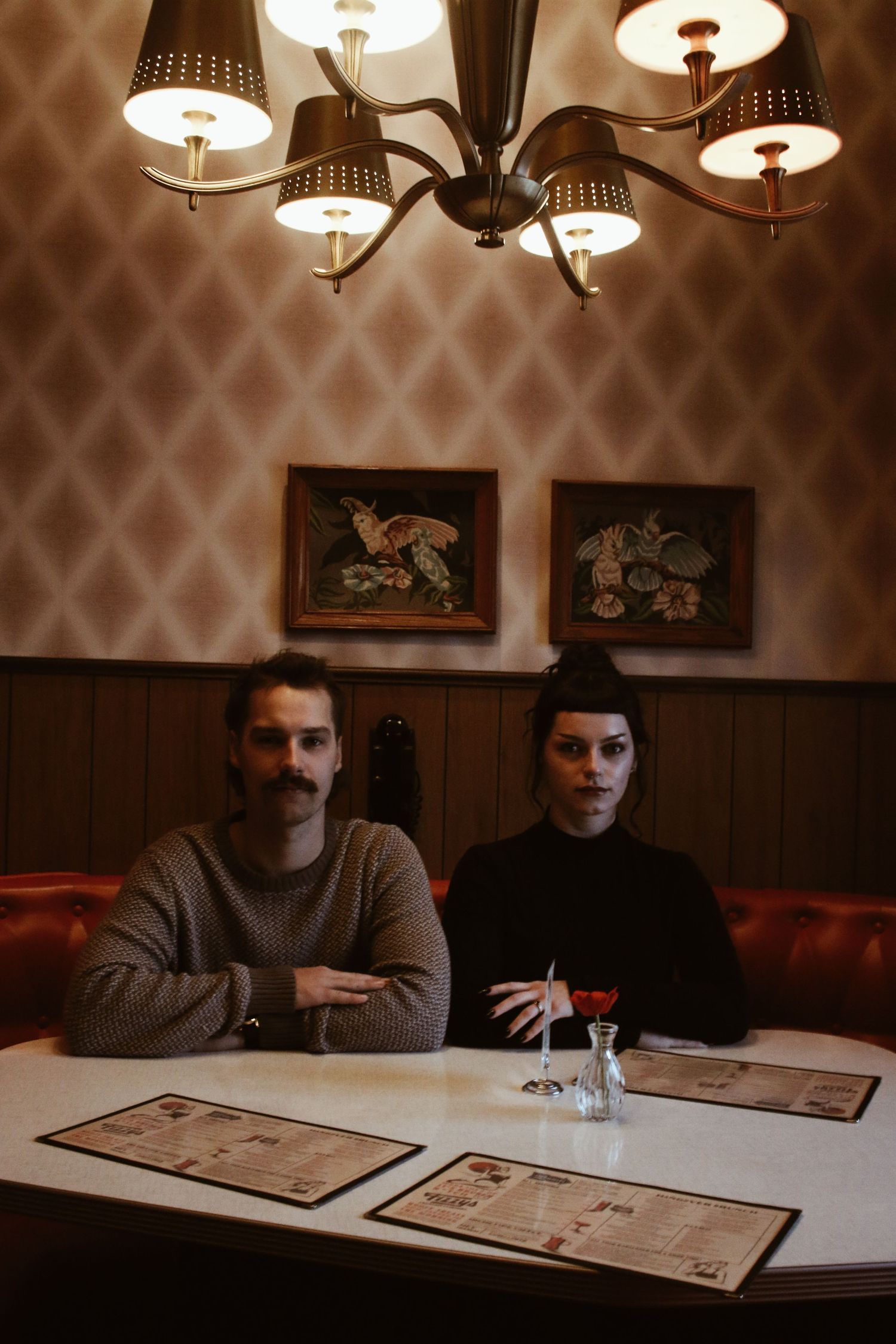 A man and woman sit in a vintage restaurant booth under a chandelier, with menus on the table and framed art behind them.