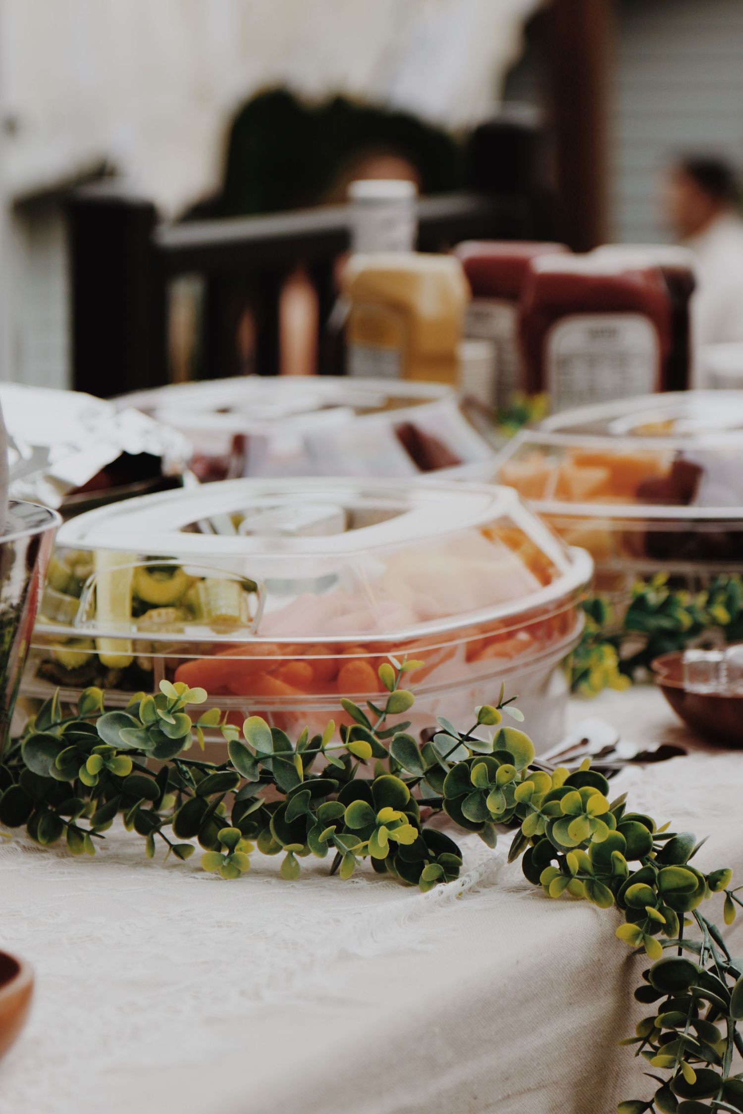 Plastic party trays with fresh vegetables and olives on a white tablecloth decorated with green eucalyptus garland.