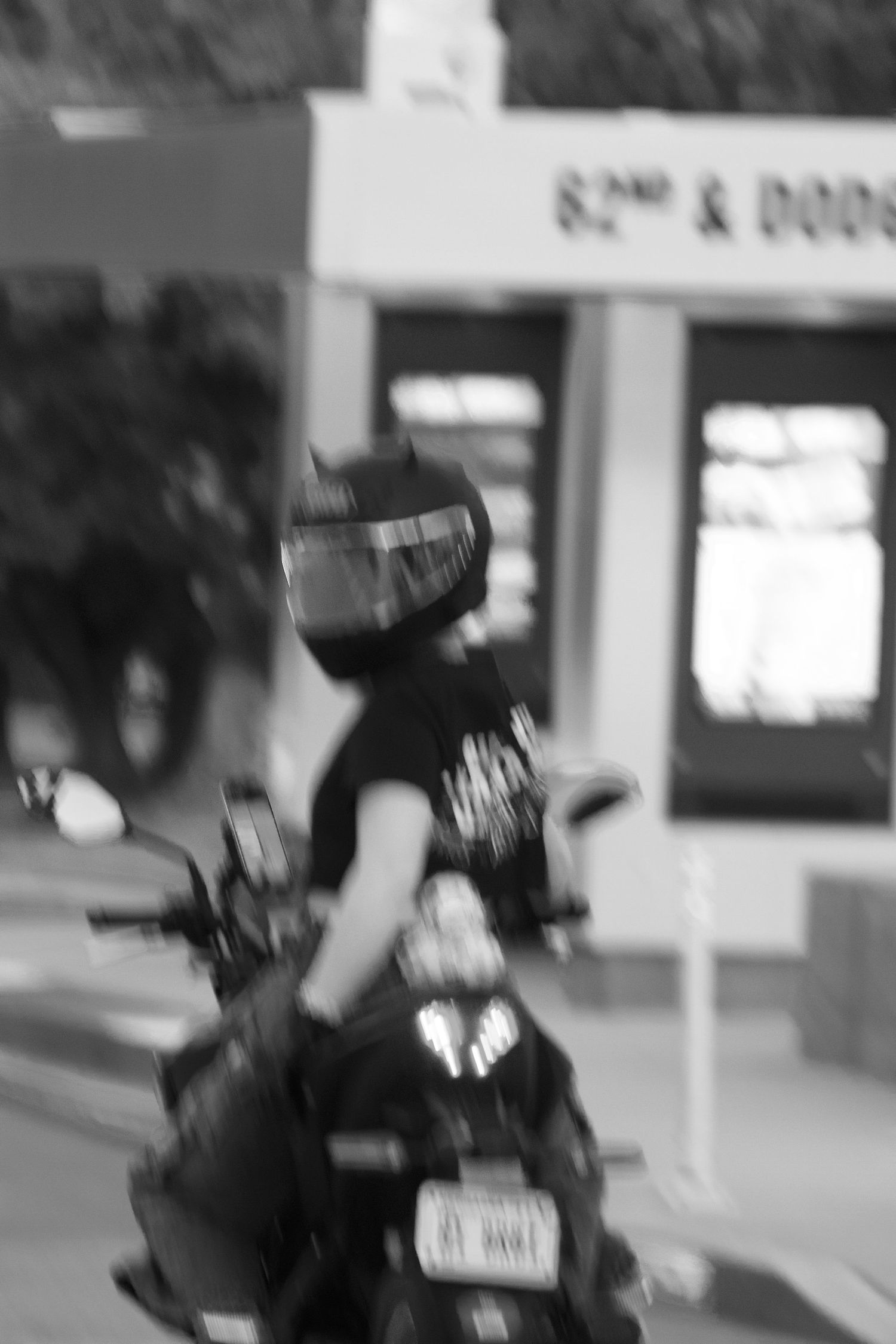 Black and white photo of a motorcyclist wearing a helmet riding past a store at 82nd and Dodge street.