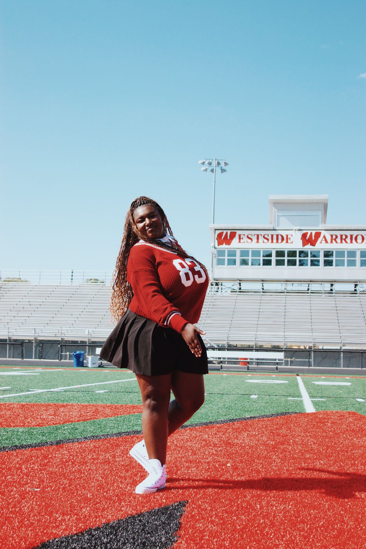 Young woman in red jersey and black skirt posing on Westside Warriors football field on a sunny day.