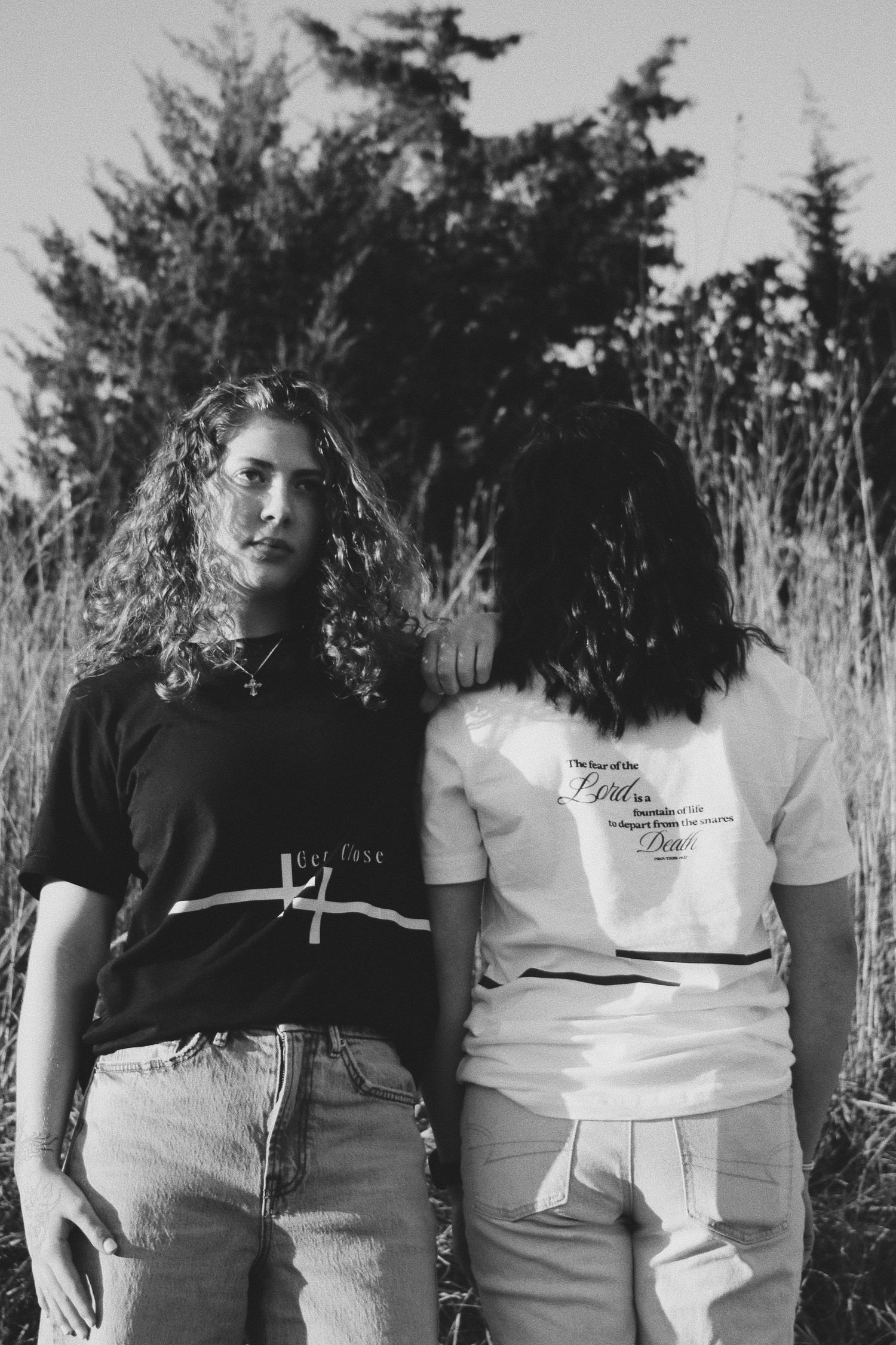 Black and white photo of two people wearing graphic tees outdoors, one facing forward and one showing the back print.