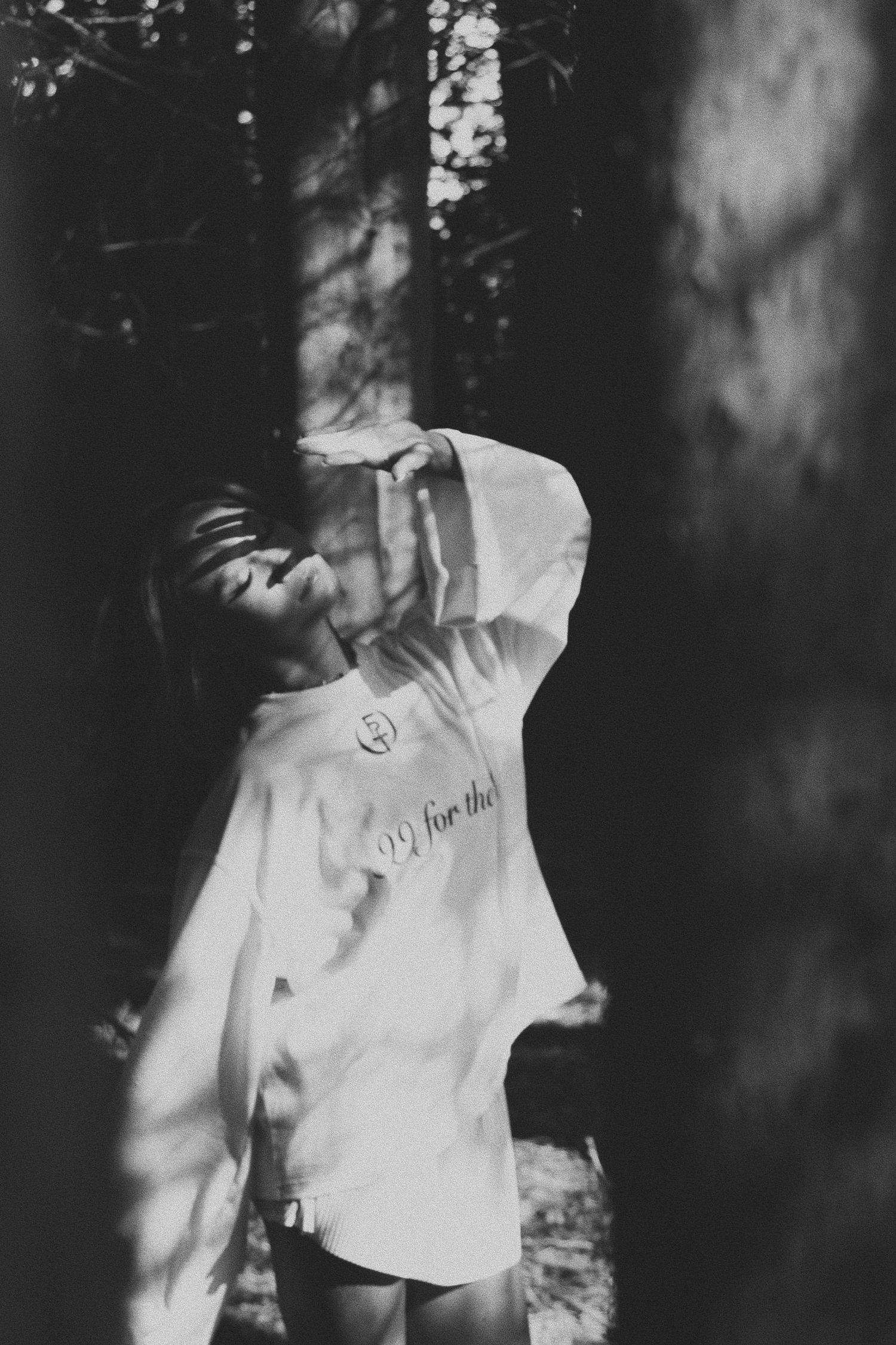 Black and white photo of a woman in an oversized white hoodie posing among trees in a forest.