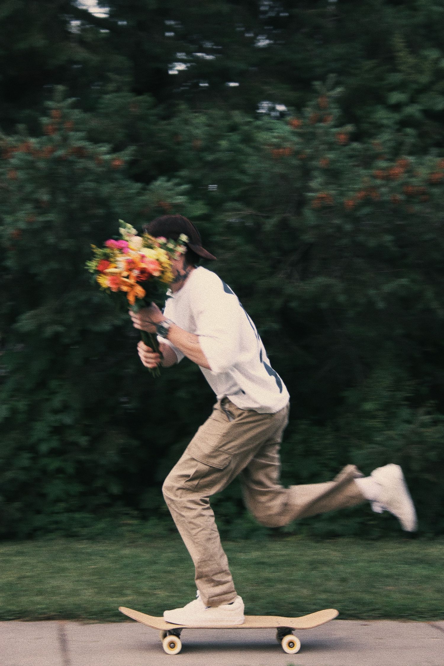 Person skateboarding while holding a colorful flower bouquet, speeding past green trees on a pathway.