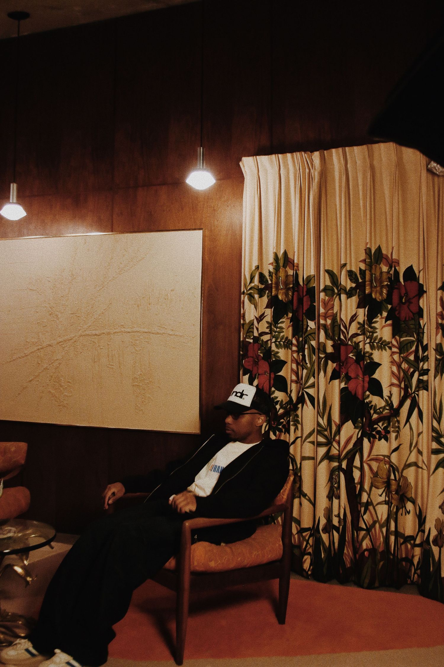 Man in cap relaxing in chair beside floral curtain and abstract art in dimly lit vintage room.
