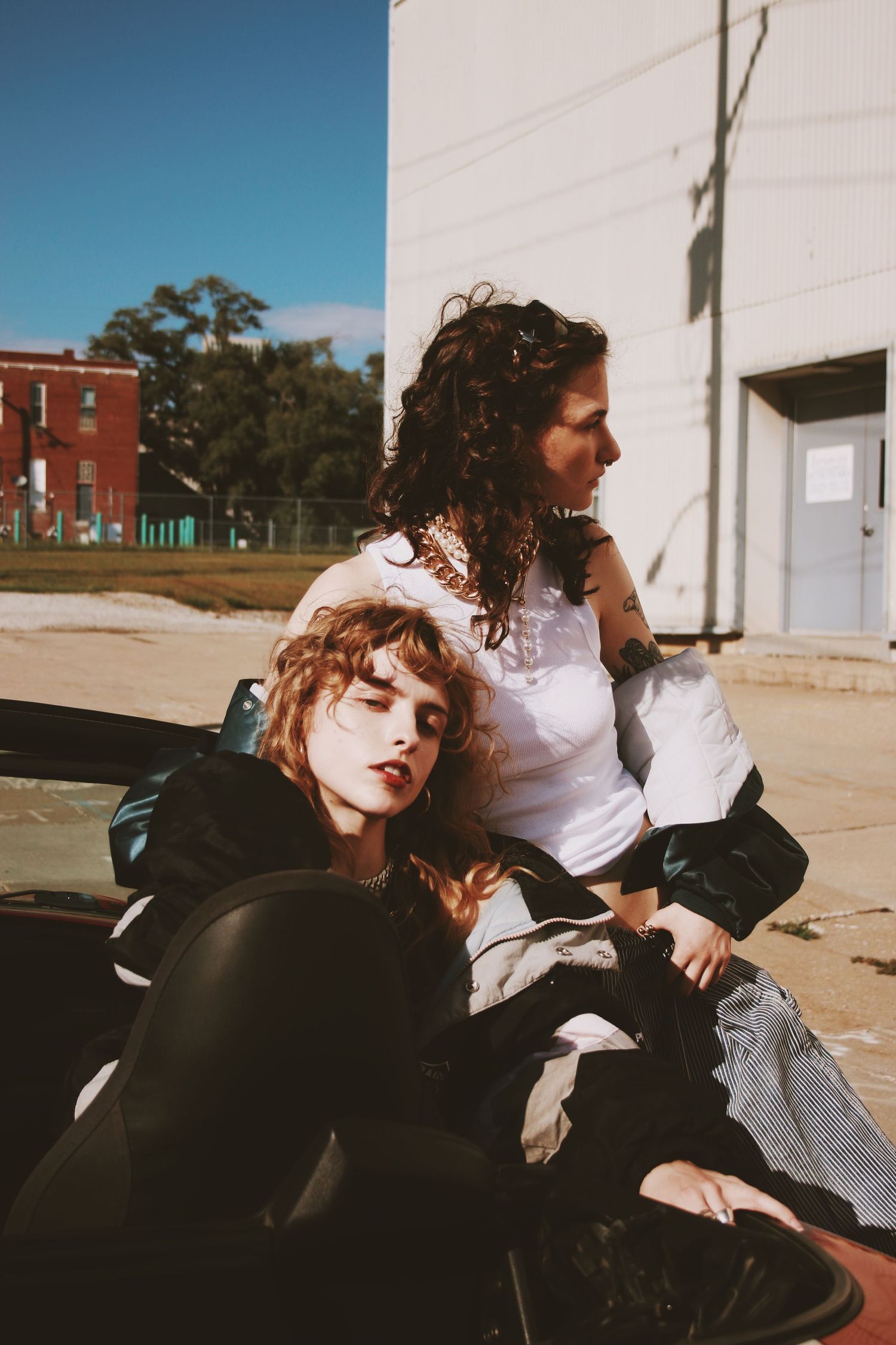 Two young women pose outdoors near a building, one reclining against the other in stylish dark and white outfits.