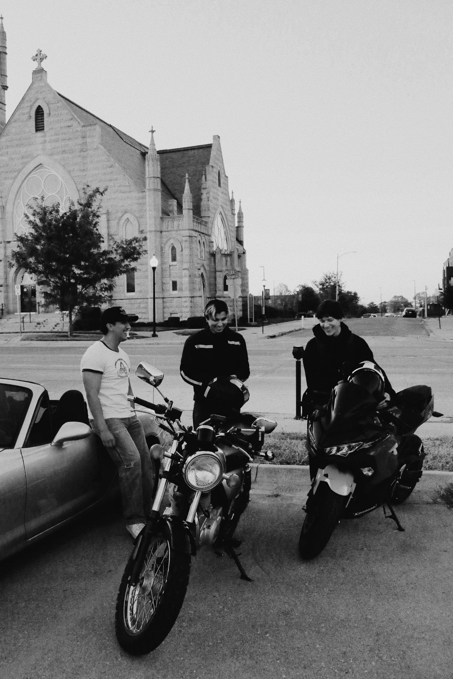 Three men with motorcycles parked outside a historic Gothic church in a black and white street photo.