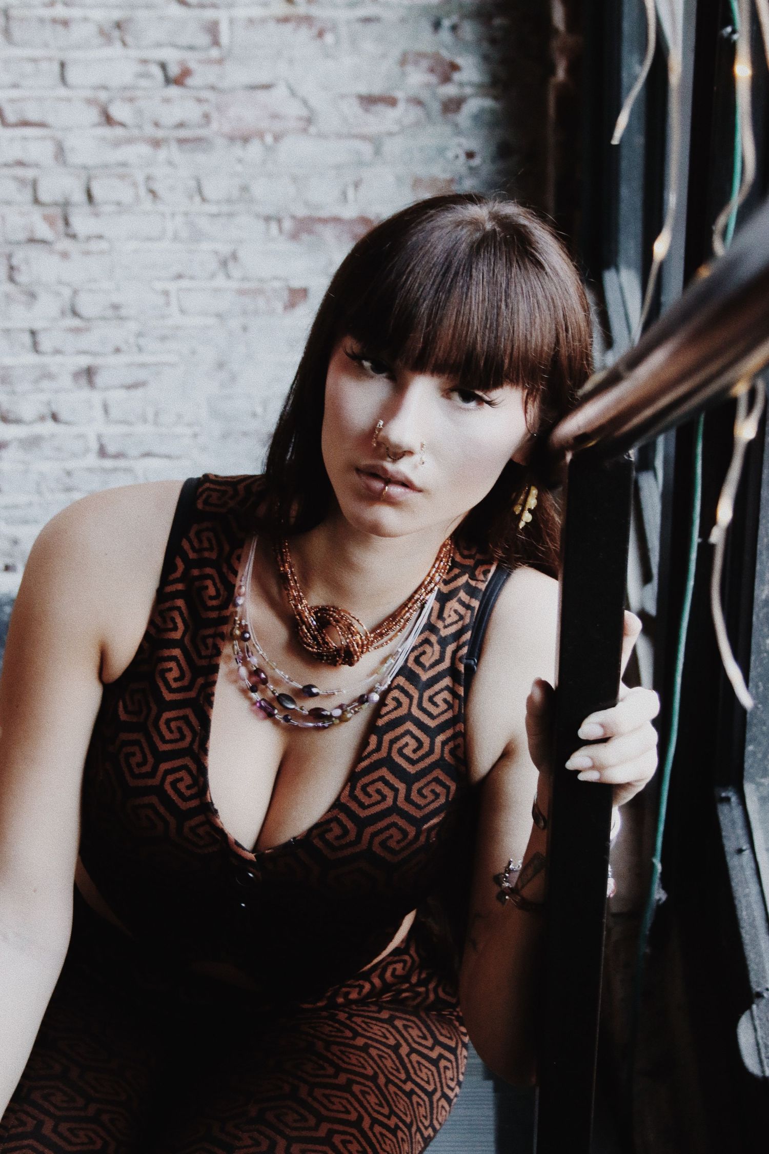 Young woman in patterned dress with layered gold necklaces posing against a white brick wall.