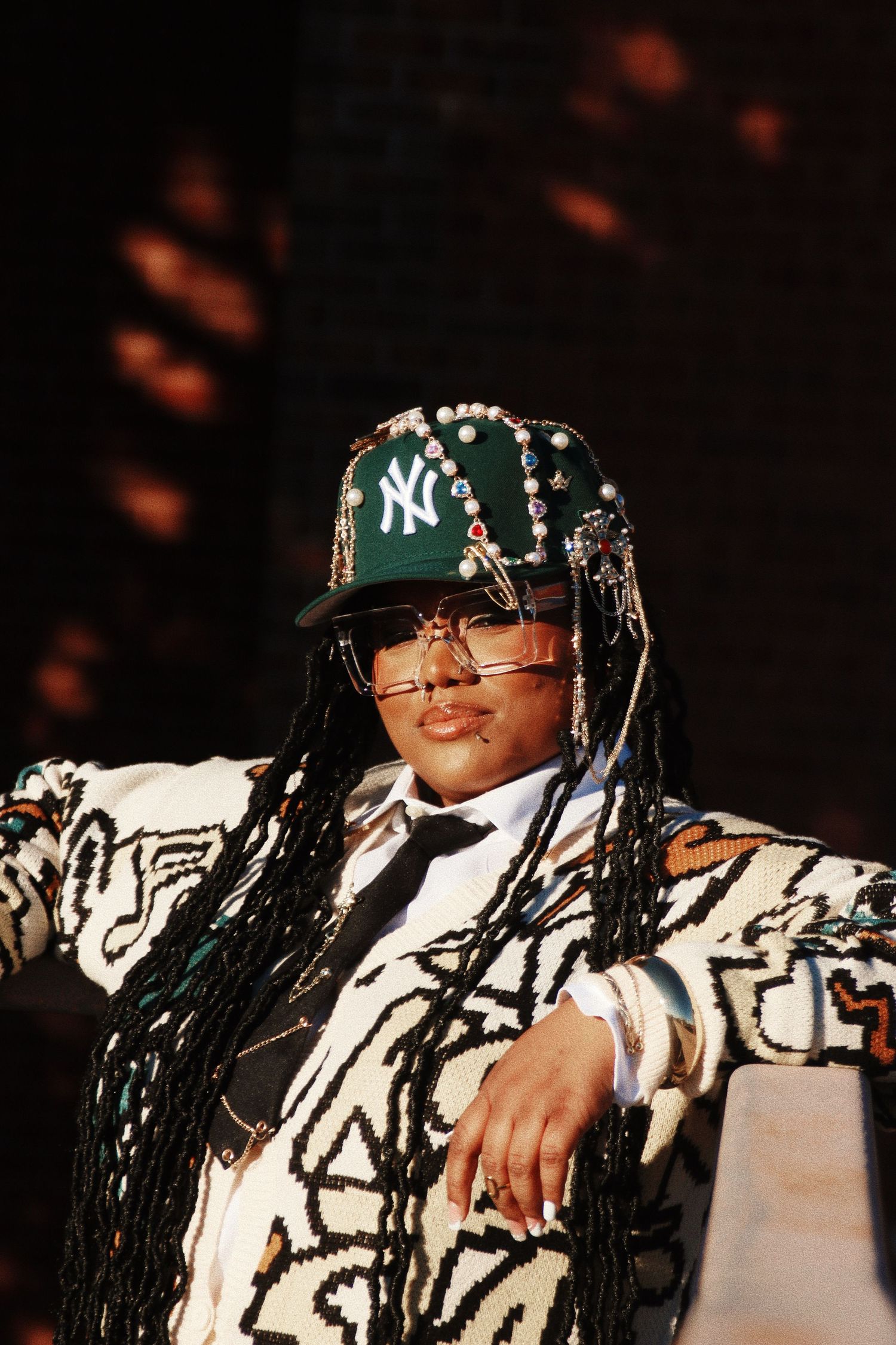 Person wearing bedazzled NY Yankees cap, long braids, glasses, and bold graffiti-print jacket posing confidently on stage.