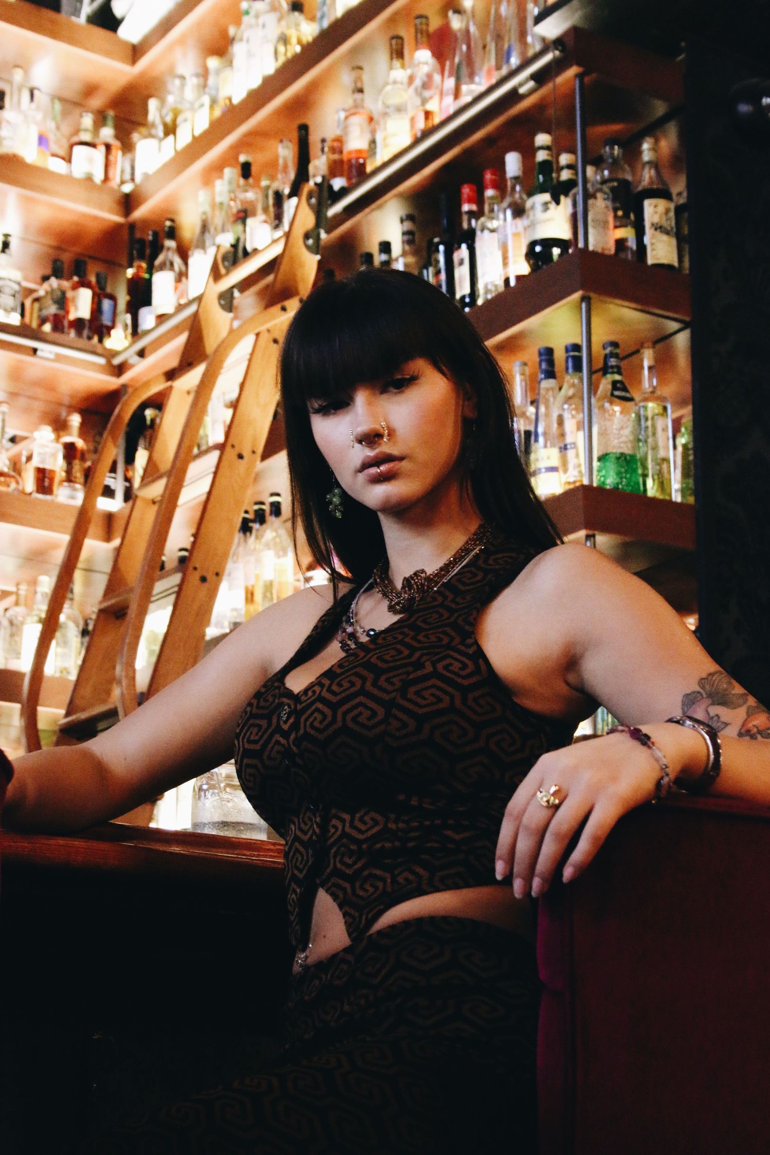 A tattooed woman in a dark patterned dress poses confidently at a well-stocked bar with shelves of liquor bottles.