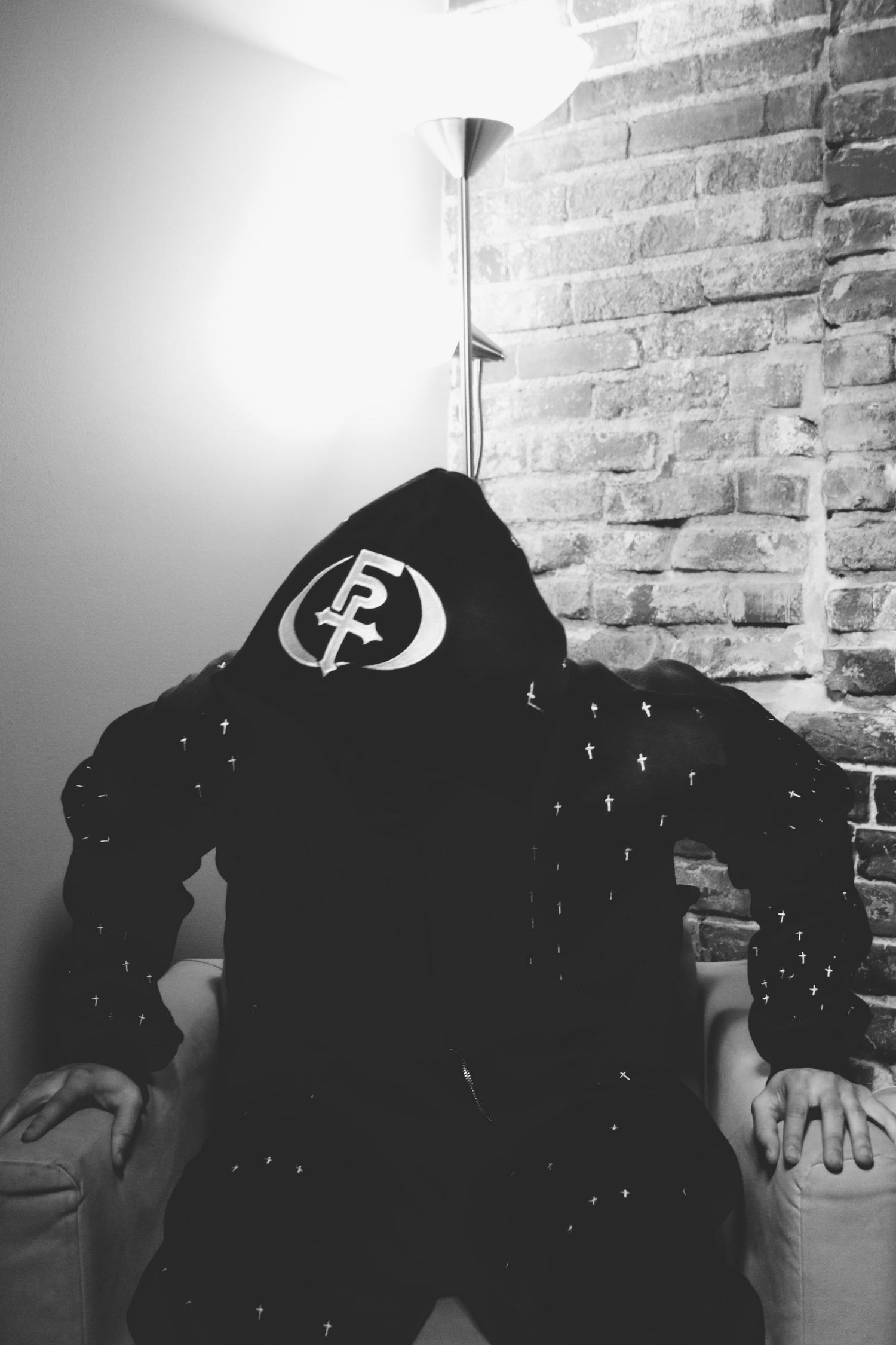 Black and white photo of person in dark hoodie with logo, sitting against brick wall with floor lamp.