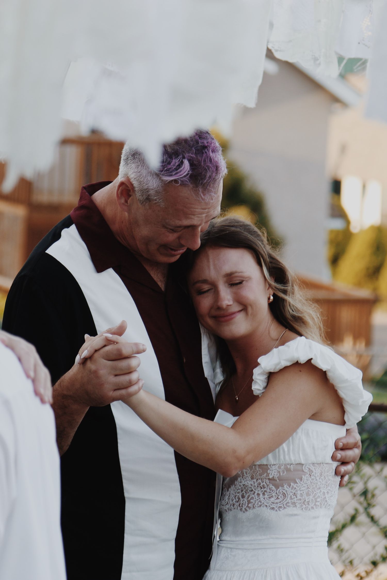 A bride in a white lace dress shares a tender first dance with a man with purple-tipped hair at an outdoor wedding.