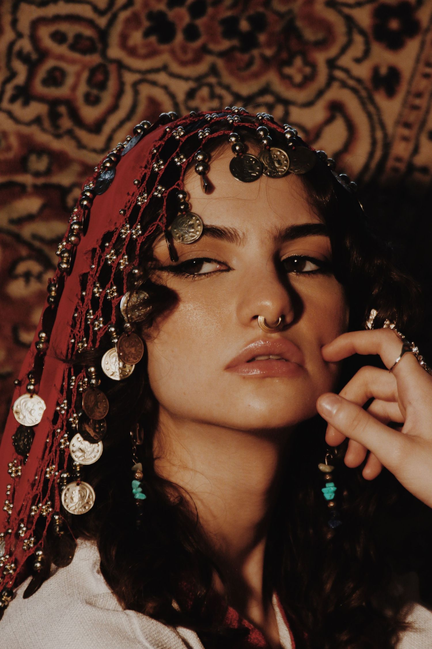 Woman wearing red coin headpiece and turquoise earrings poses against ornate tapestry background.