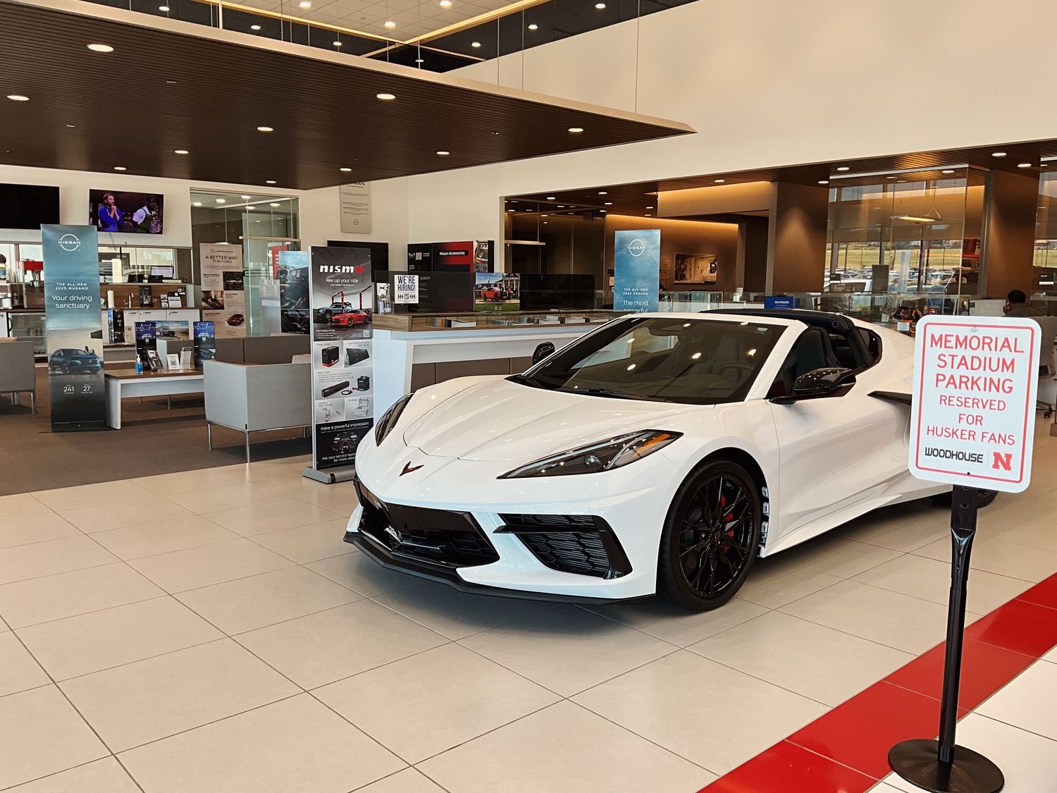 White Chevrolet Corvette C8 displayed in a bright car dealership showroom with red velvet rope barriers.