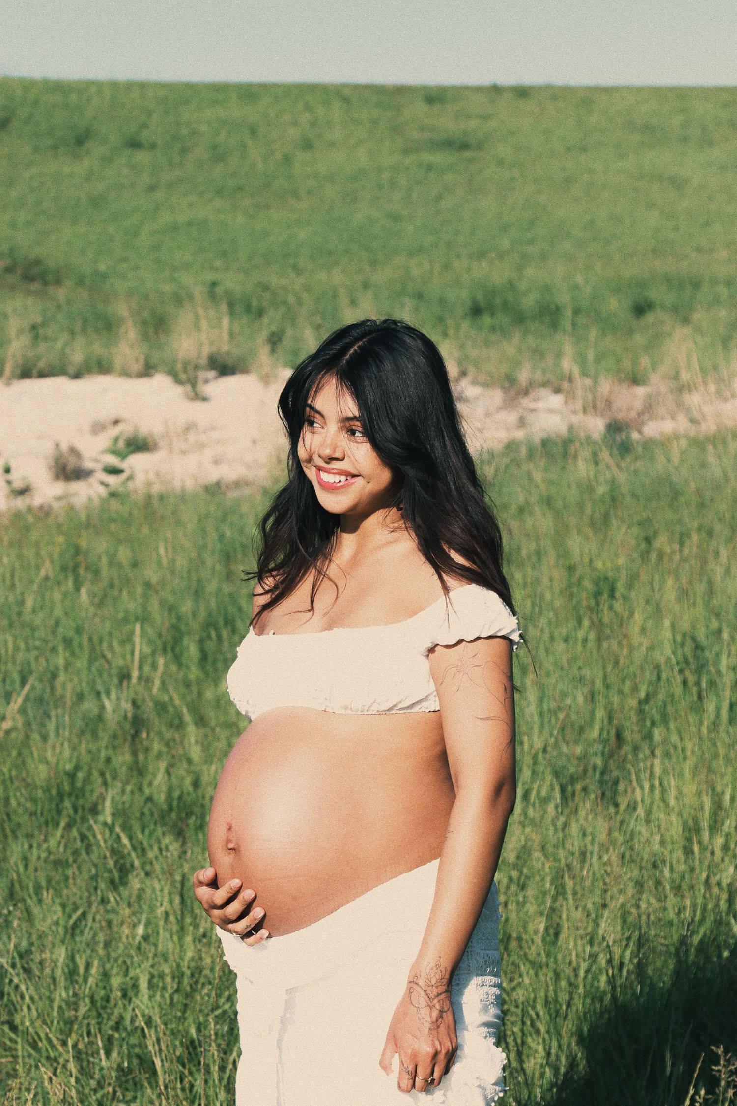 Smiling pregnant woman in white crop top holding baby bump outdoors in green field on sunny day.