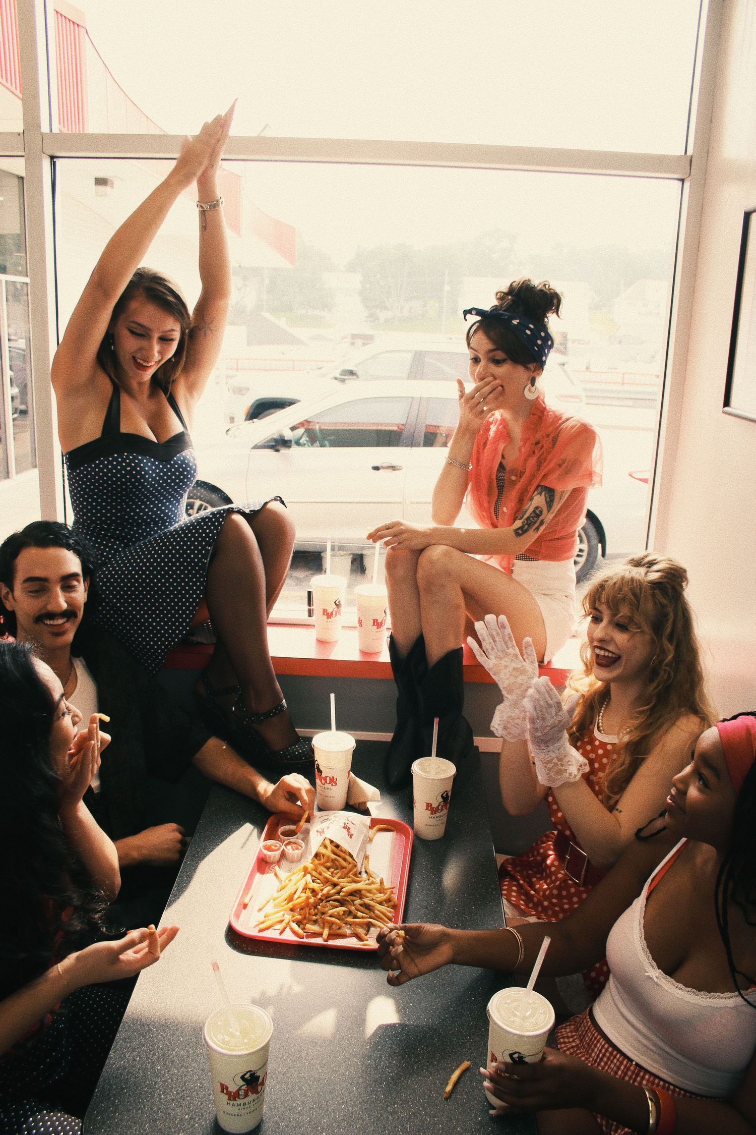 Group of retro-styled friends sharing fries and drinks at a fast food restaurant booth, laughing and celebrating together.