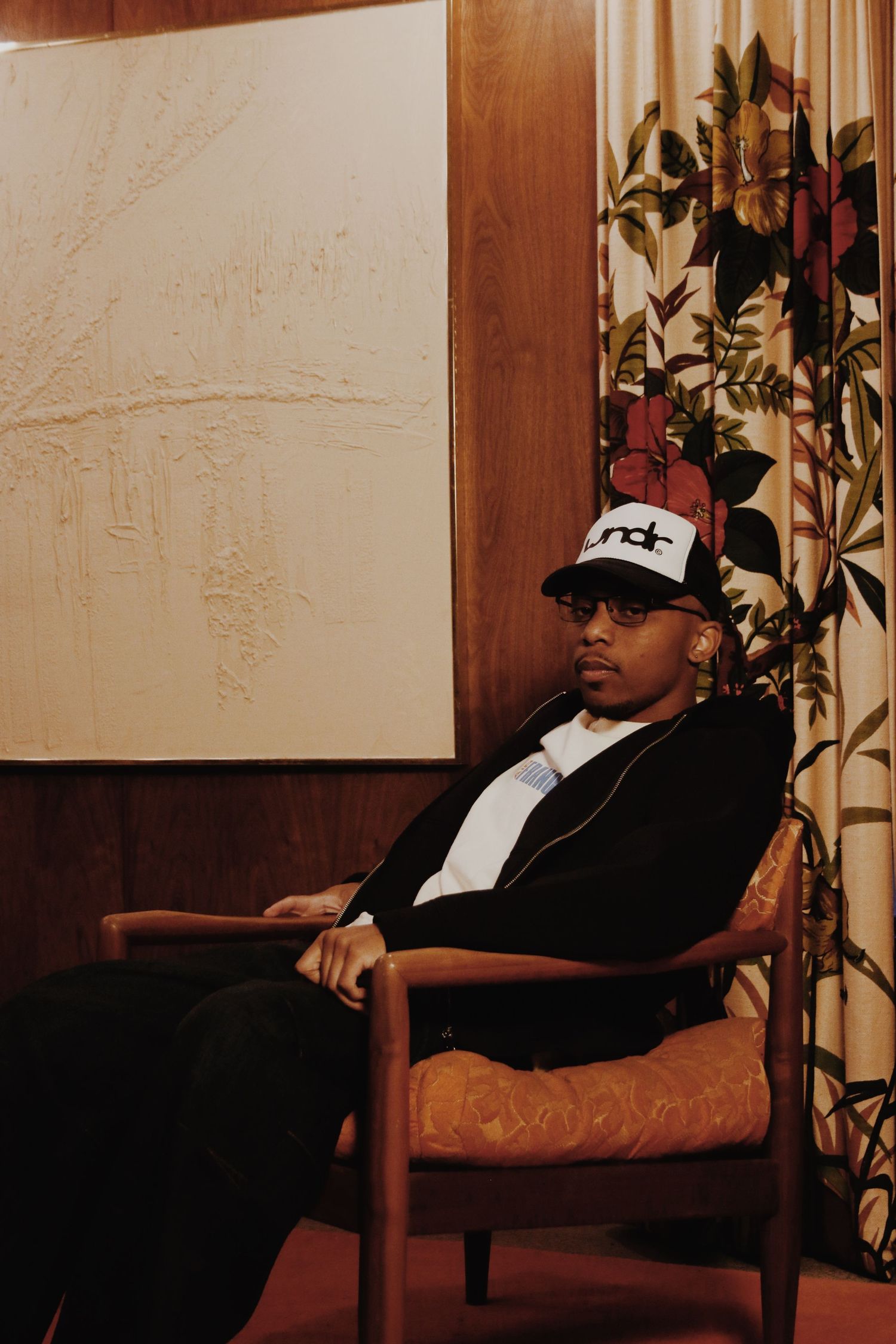 Man in black jacket and snapback cap sitting relaxed in a wooden chair near floral curtains and abstract wall art.