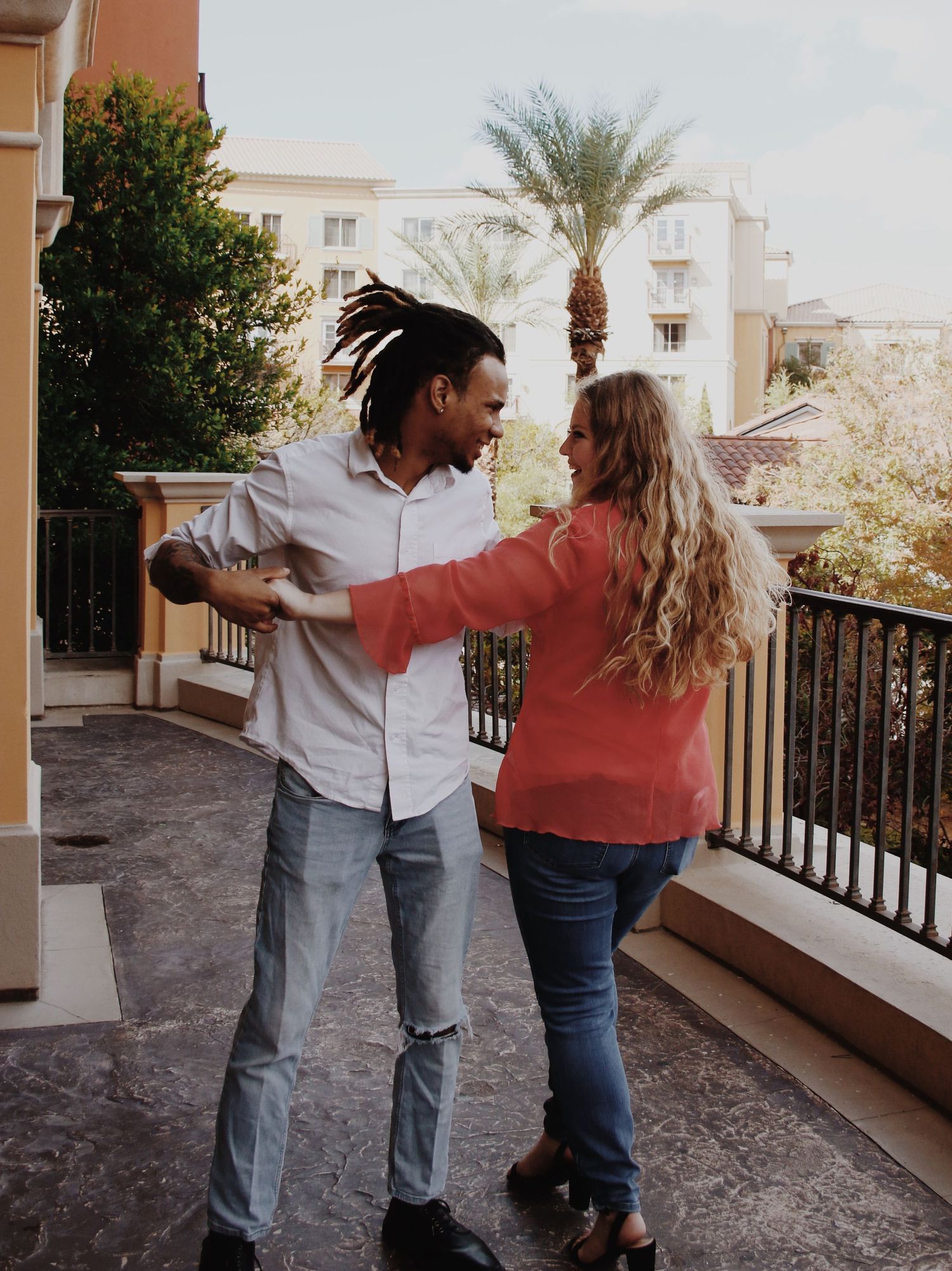 A couple dances joyfully on a balcony, surrounded by palm trees and buildings in a sunny urban setting.
