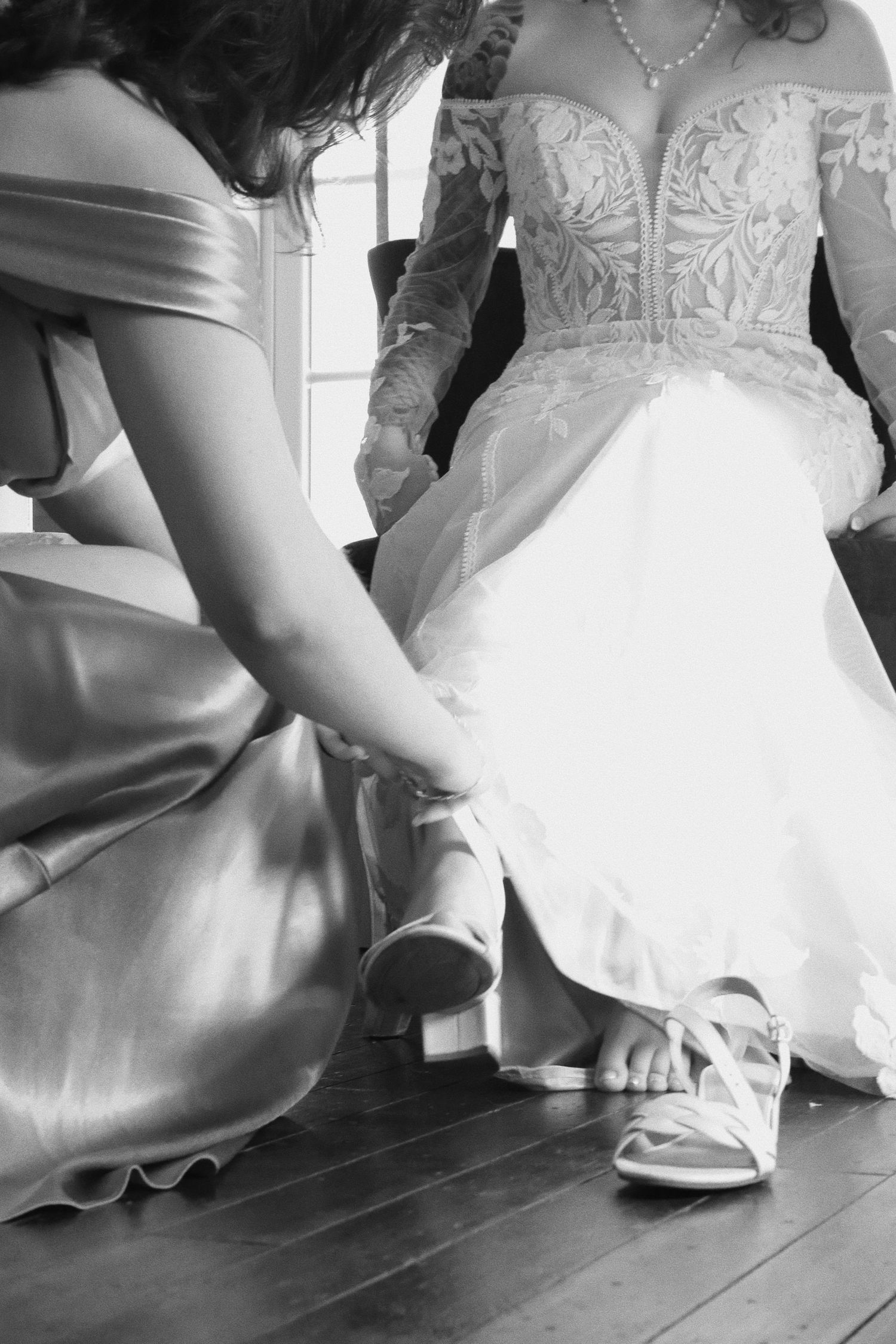 Bridesmaid in satin gown helping bride put on wedding heels, black and white photo.
