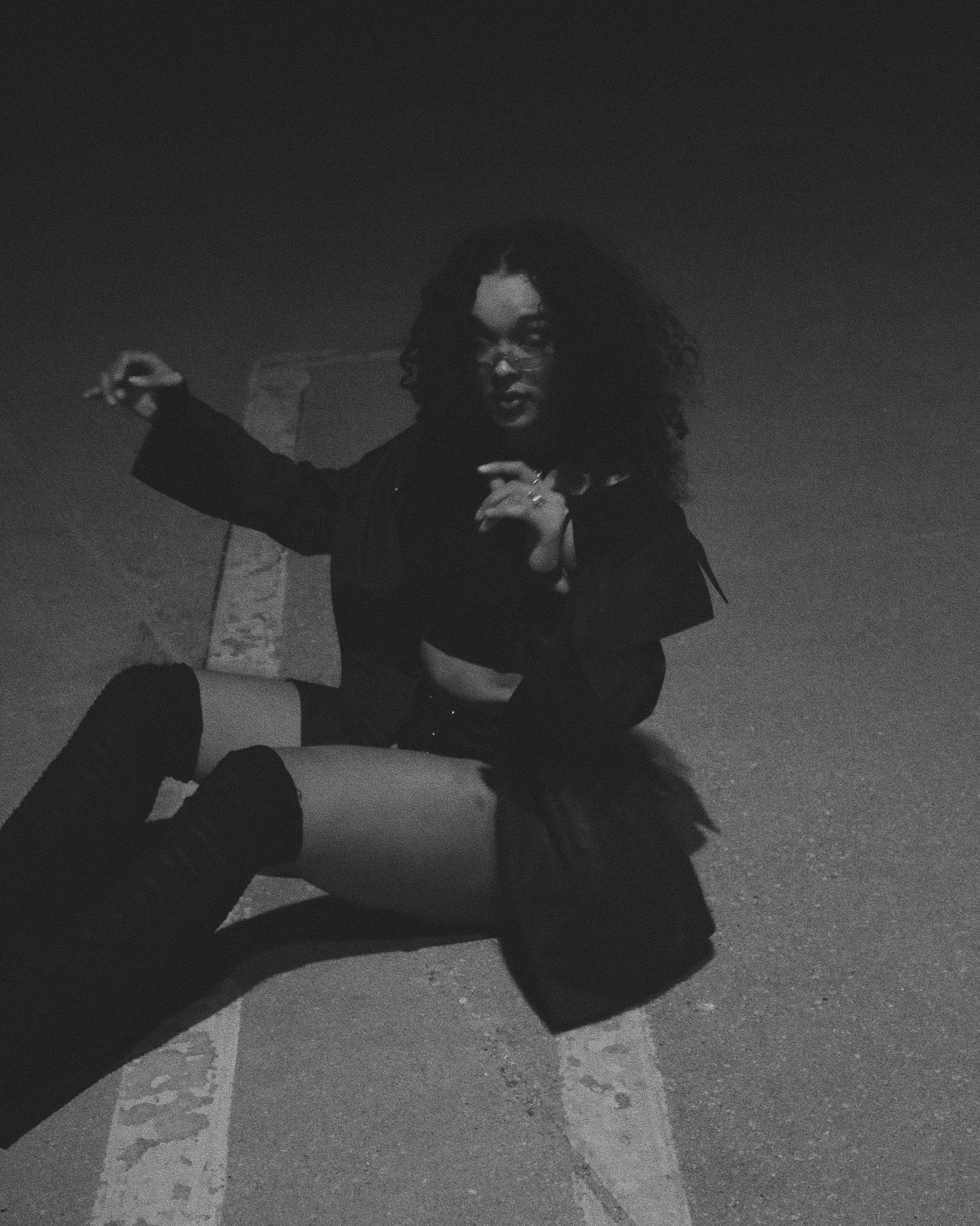 Black and white photo of a young woman with curly hair sitting on pavement, wearing knee-high boots and a crop top.