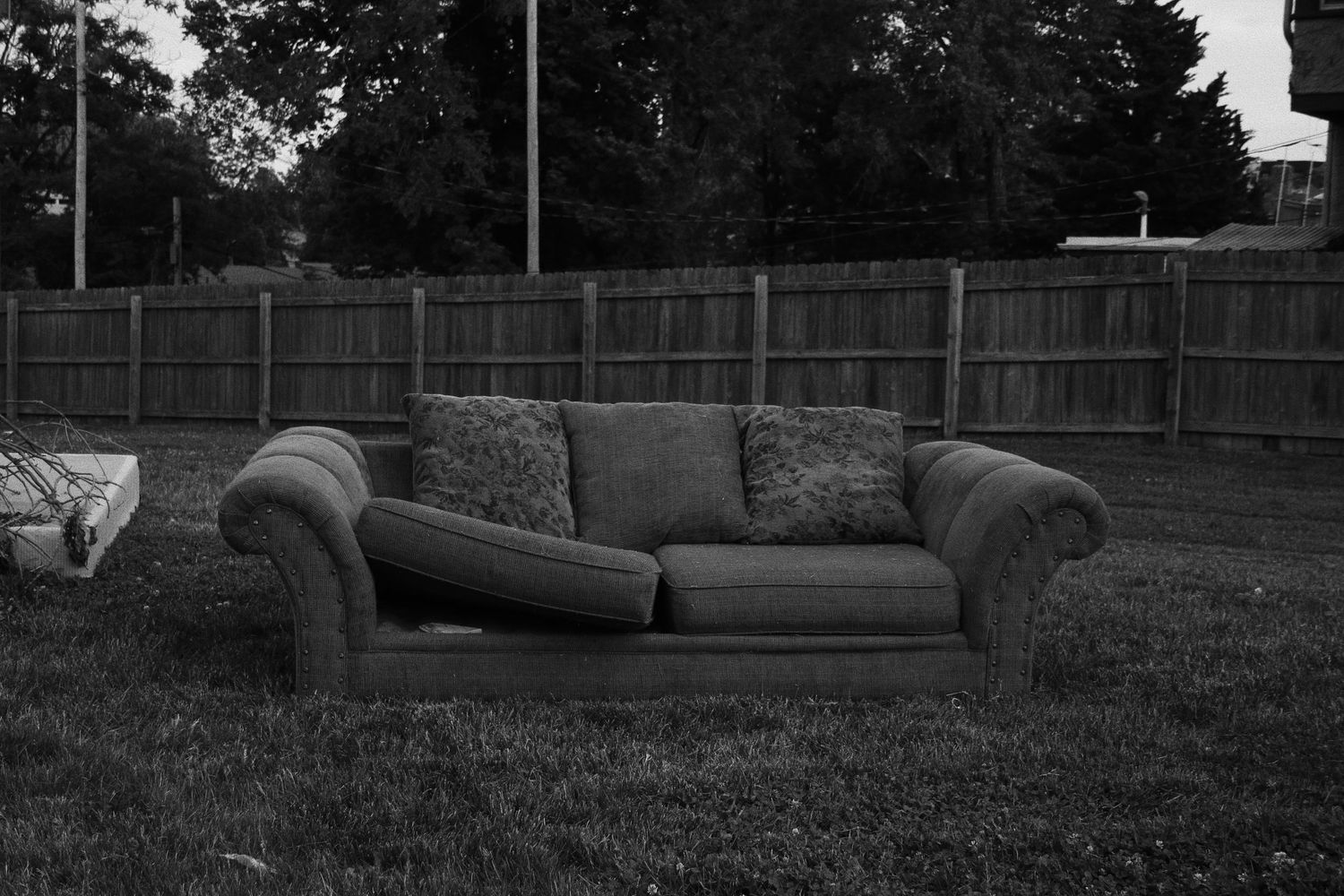 Black and white photo of an old sofa with pillows sitting abandoned on a grass lawn near a wooden fence.