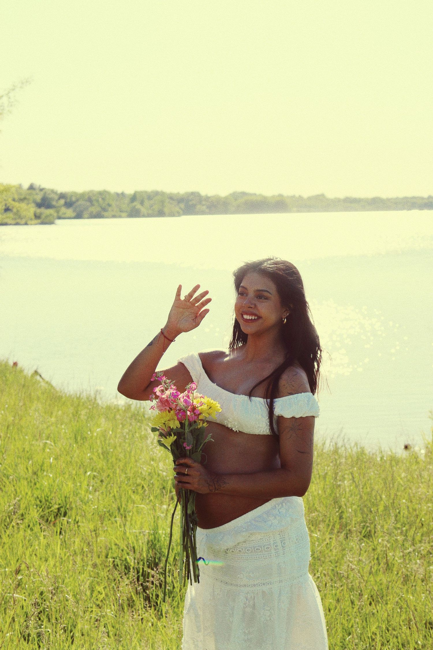 Smiling woman in white crop top holding colorful flowers, waving near a sunny lakeside with green grass.