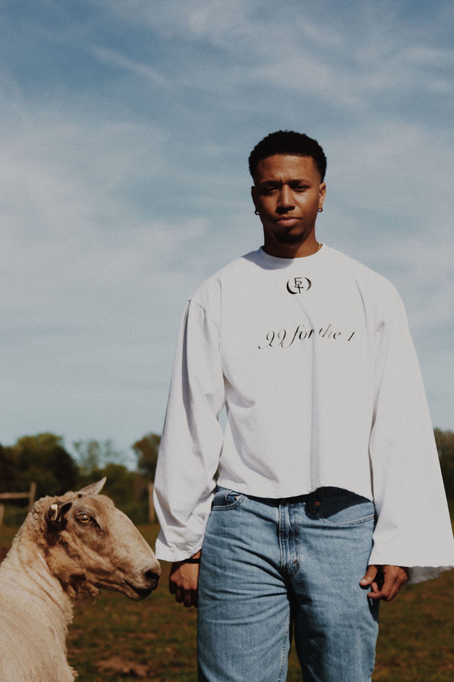 Young man in white graphic long sleeve shirt and jeans stands beside a sheep in an open field.