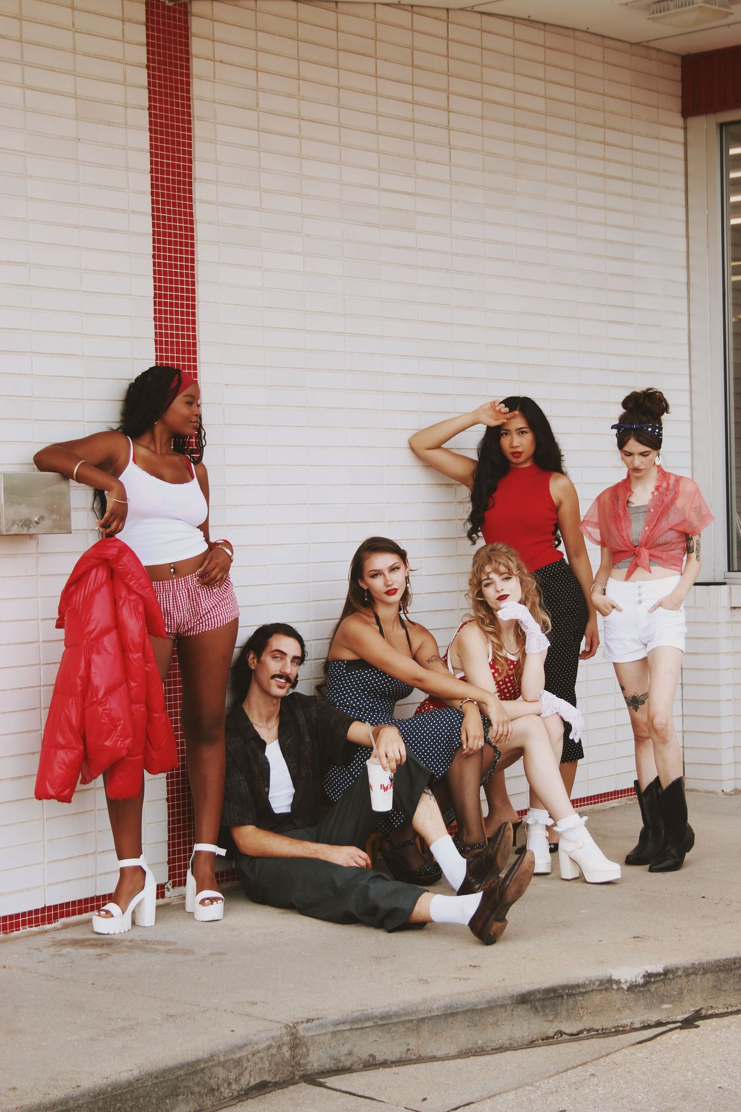 Diverse group of seven stylish young people posing against white brick wall in trendy outfits.