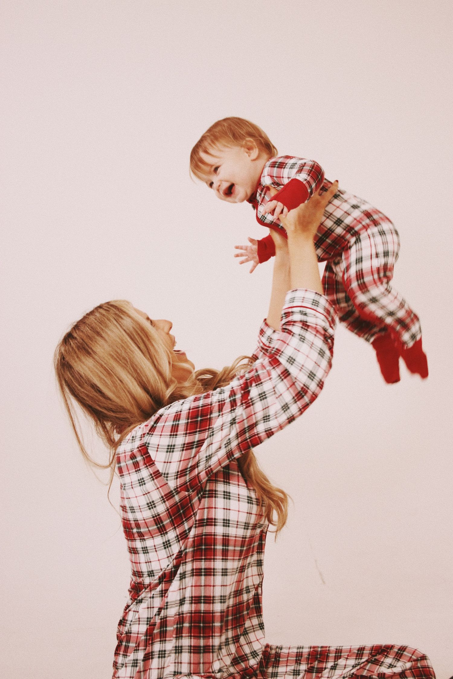 Mother and baby in matching red plaid pajamas, mom lifting laughing infant overhead on white background.