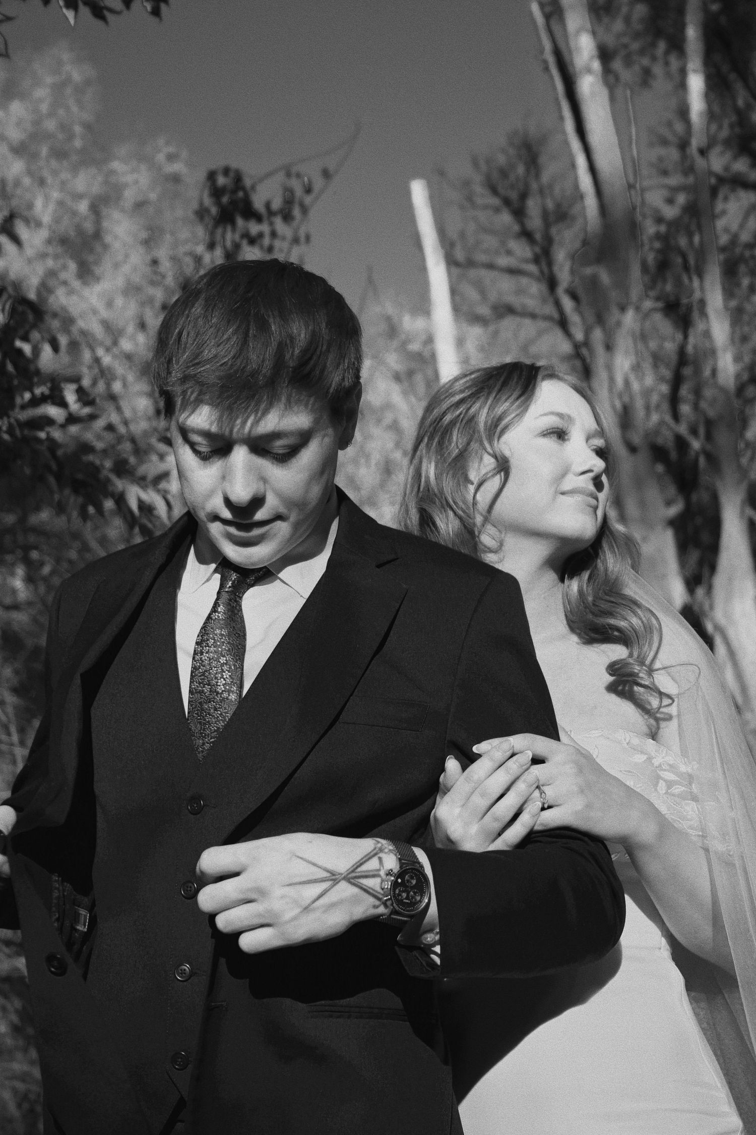 Black and white wedding photo of bride and groom standing outdoors among trees, bride resting hand on groom's arm.