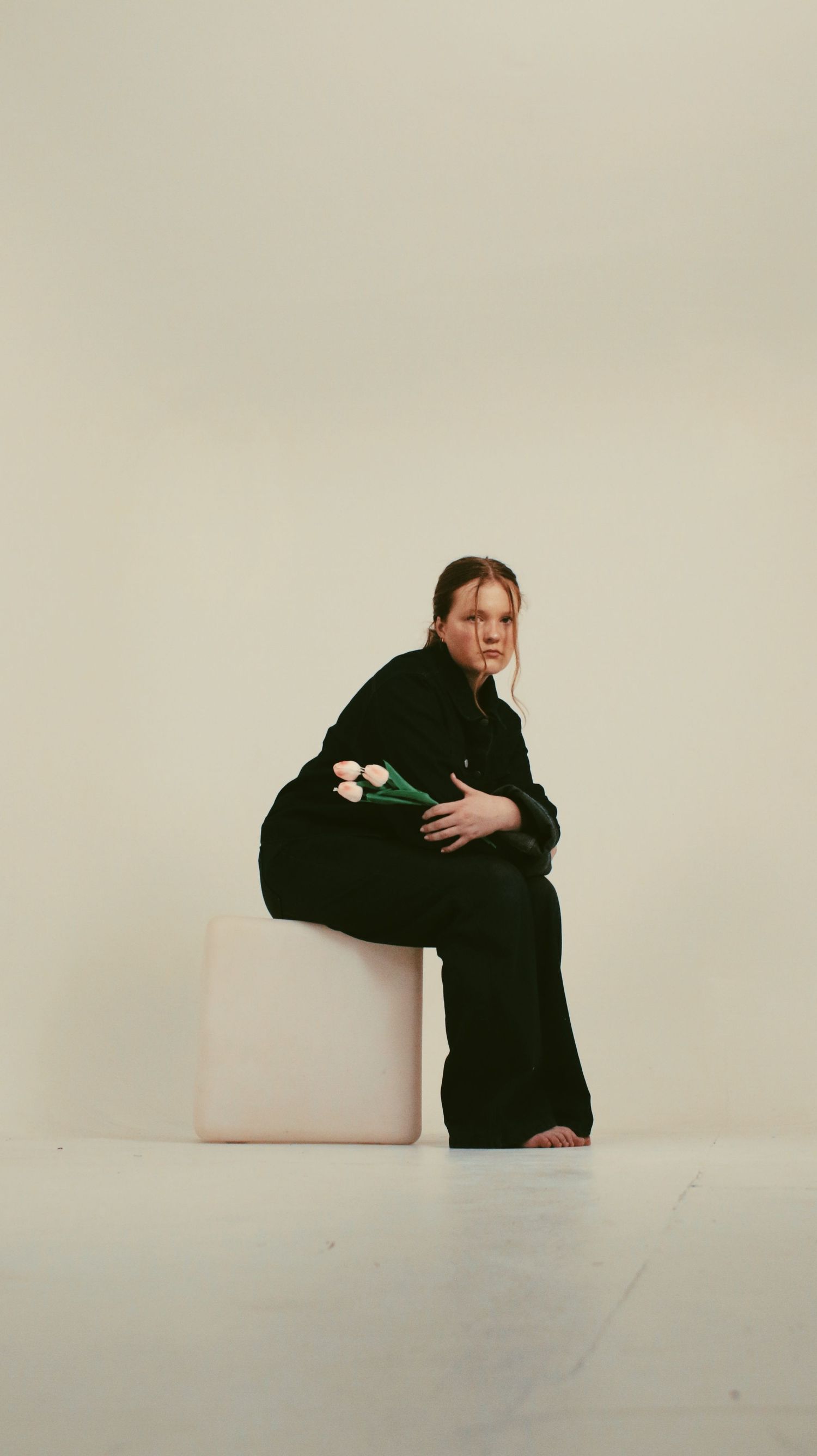 Woman in black outfit sits on white cube holding tulips against a light background.