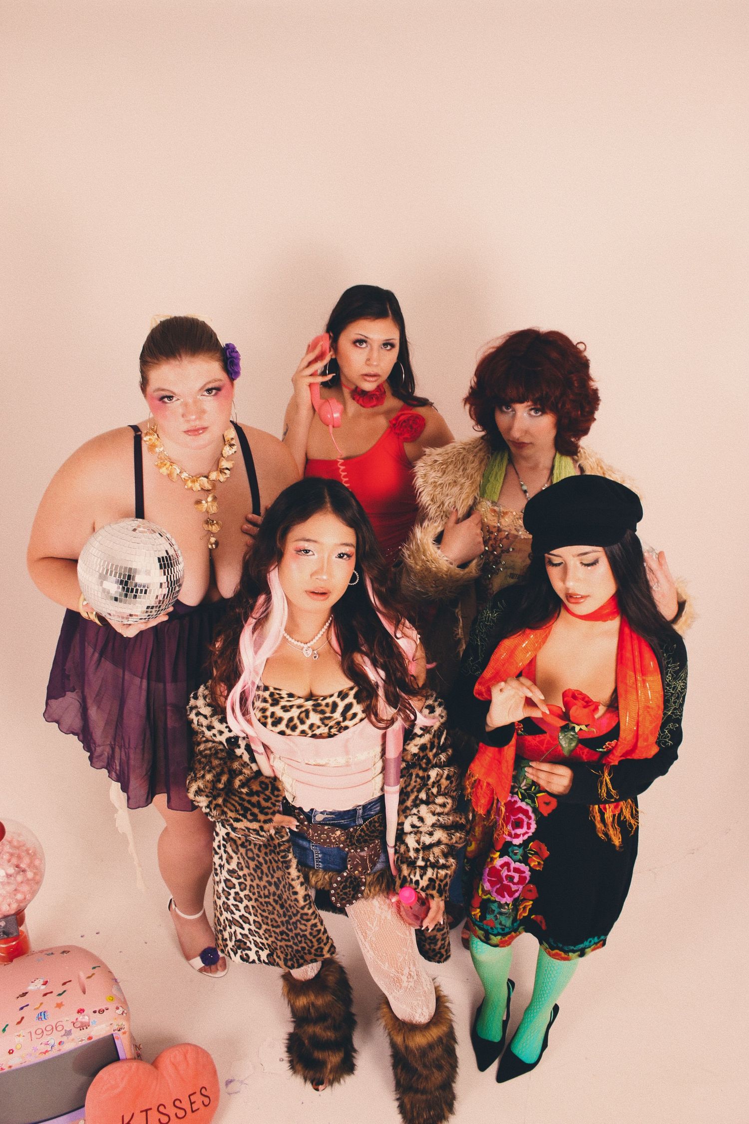 Five women in colorful, eclectic outfits pose together in a studio, one holding a disco ball.