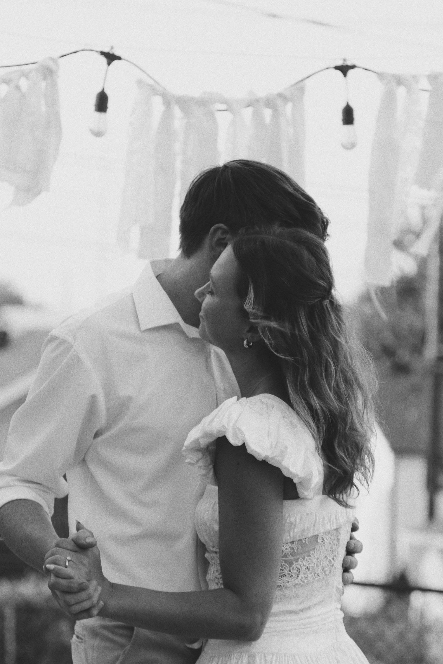 Black and white photo of a couple sharing a romantic dance at an outdoor wedding reception under string lights.