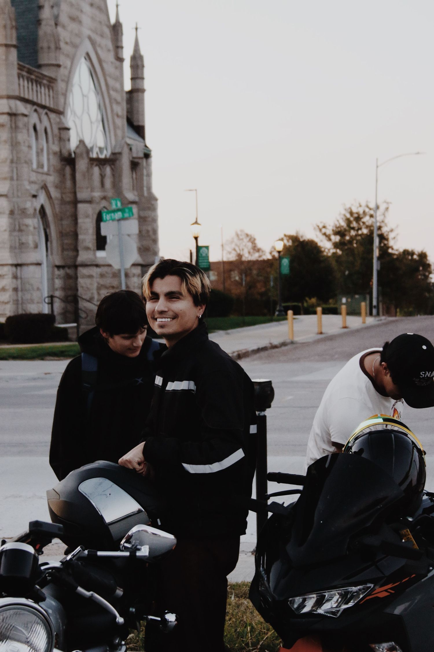 Three young men in black jackets stand near motorcycles on a street corner with a stone church in the background.