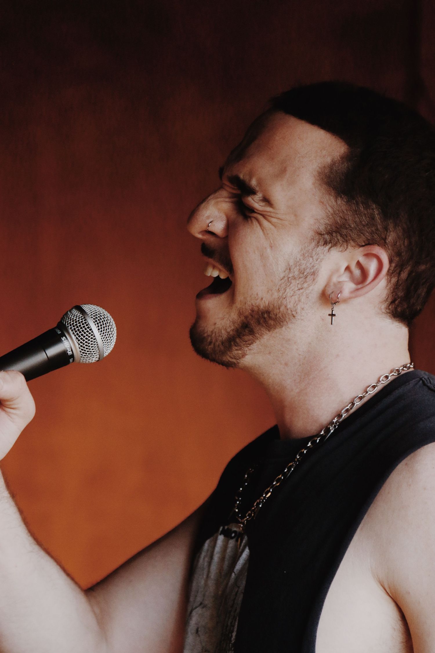 Male rock singer performing passionately into microphone, wearing black sleeveless shirt and cross earring.