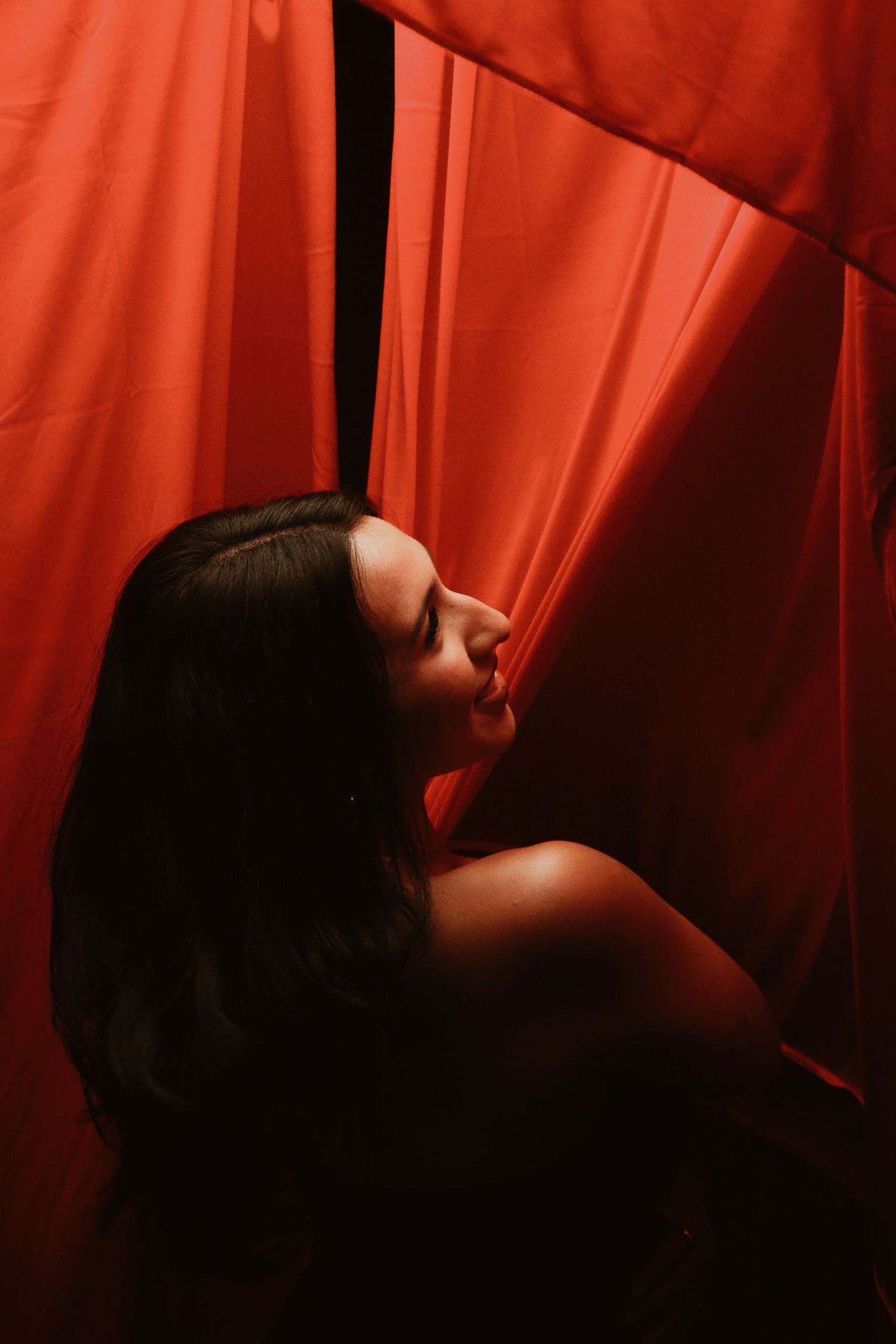A woman with dark hair poses dramatically against flowing red curtains in moody, atmospheric lighting.