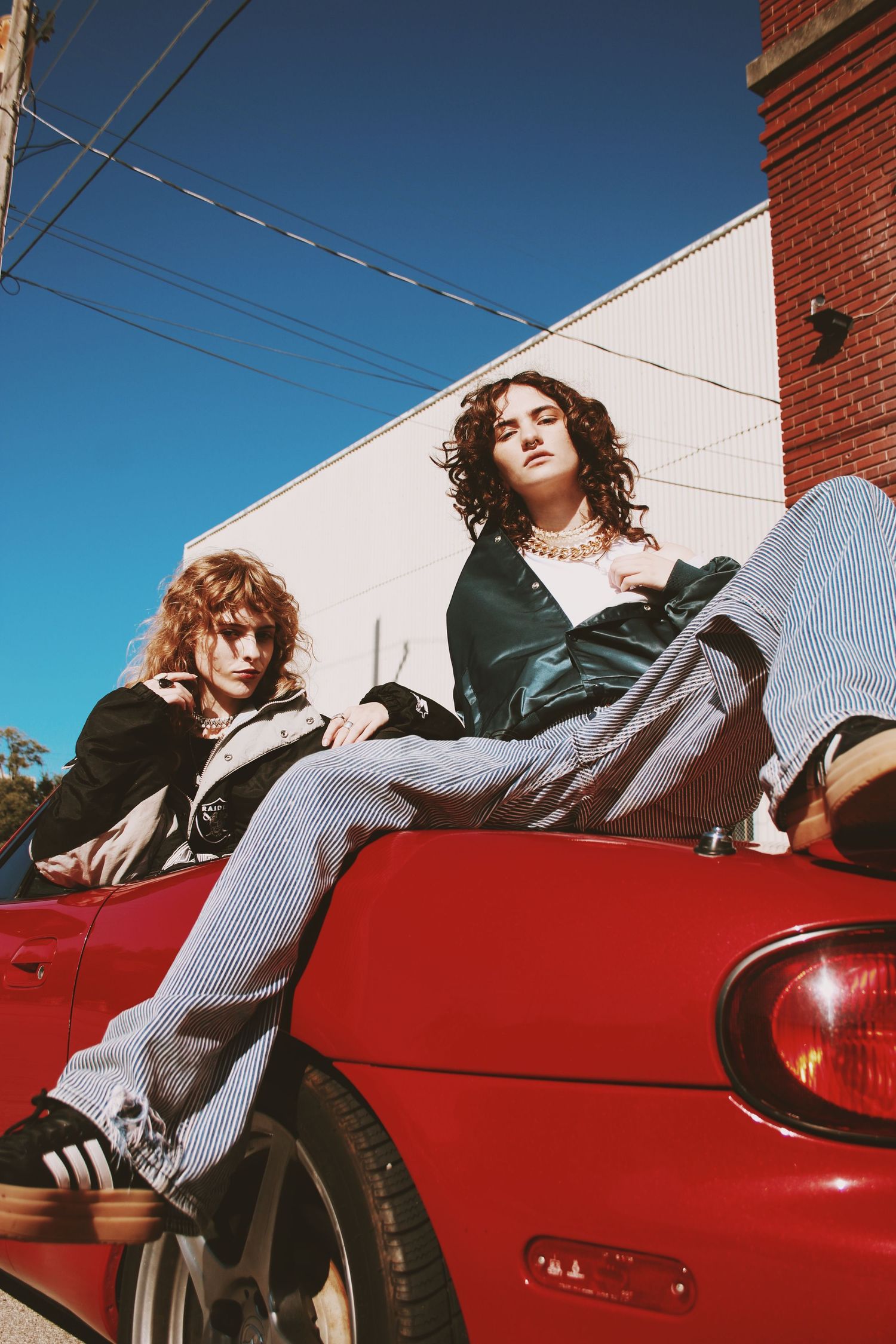 Two young people posing on a red convertible car against a blue sky and brick building backdrop.