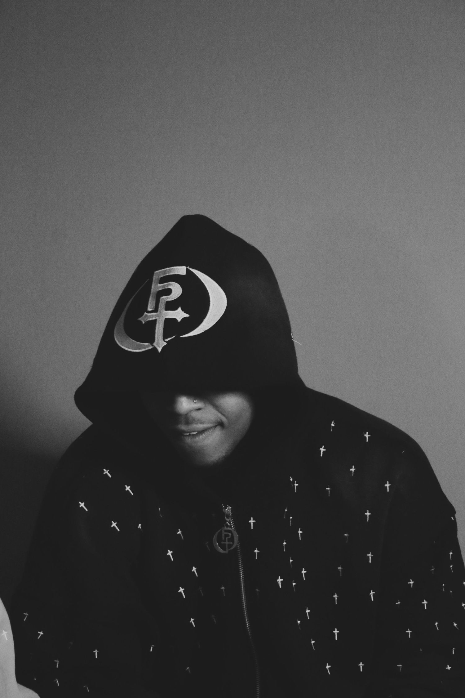 Person in black hoodie with cross pattern and logo, black and white portrait photography.