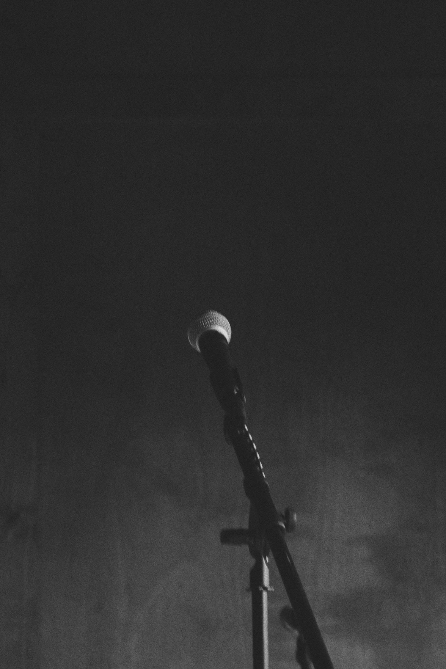 Black and white photo of a microphone on a stand against a dark background, evoking a moody performance atmosphere.