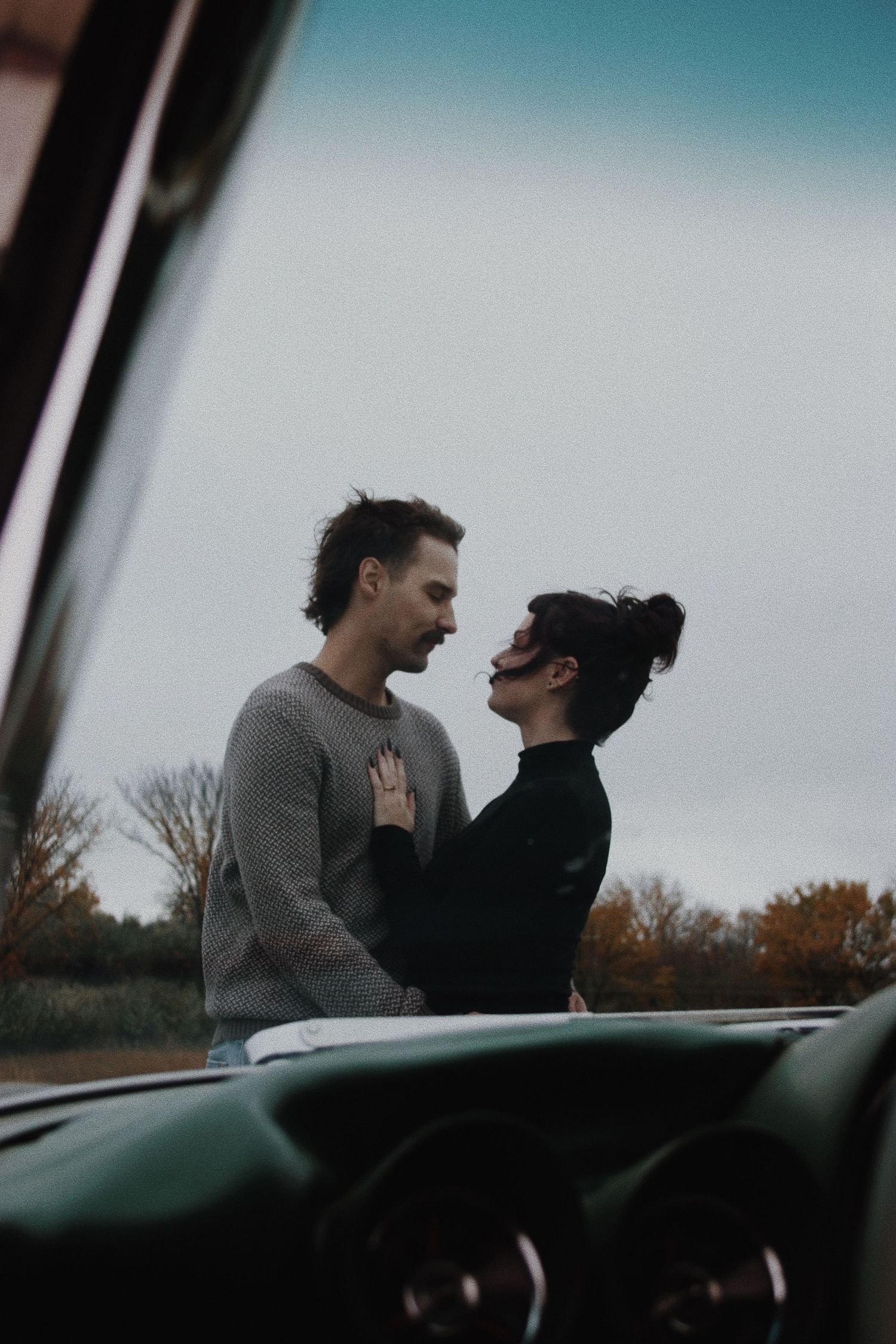 A romantic couple sharing an intimate moment standing outside a vintage car on a cloudy autumn day.
