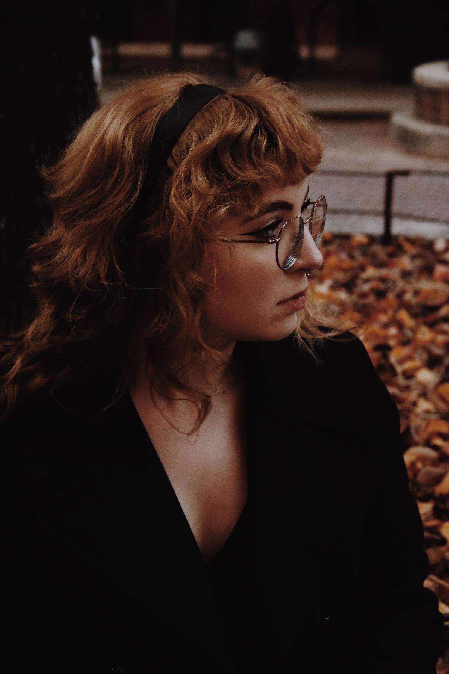 Young woman with curly red hair, black headband, round glasses, and black blazer sitting outdoors among autumn leaves.