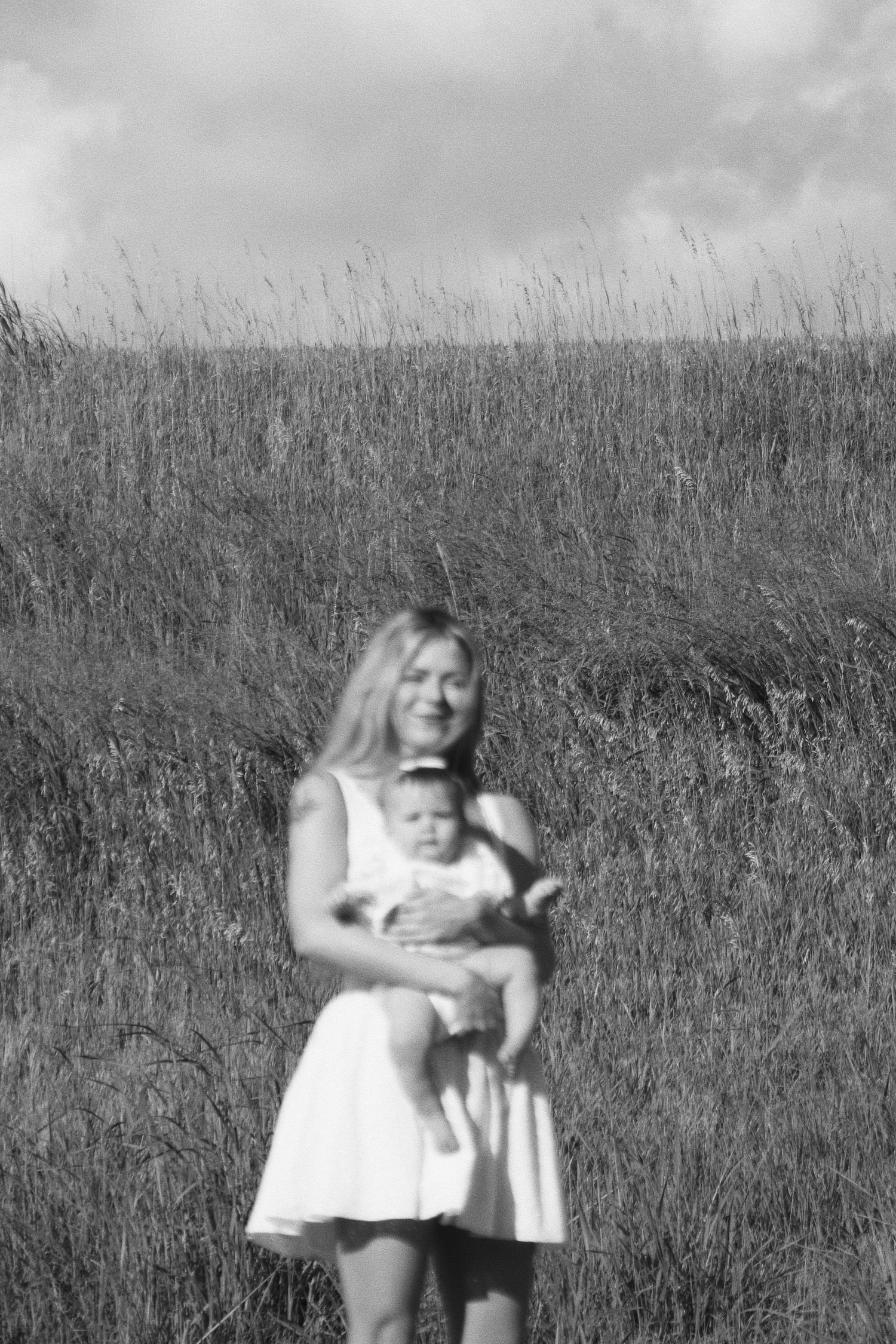 Black and white photo of a smiling blonde woman in a white dress holding a baby in a grassy field.