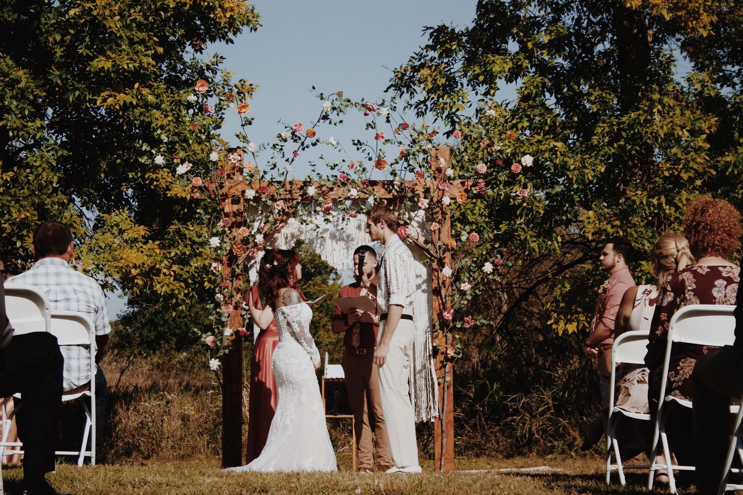 Two brides exchange vows under a floral arch at an outdoor fall wedding ceremony with white chairs.